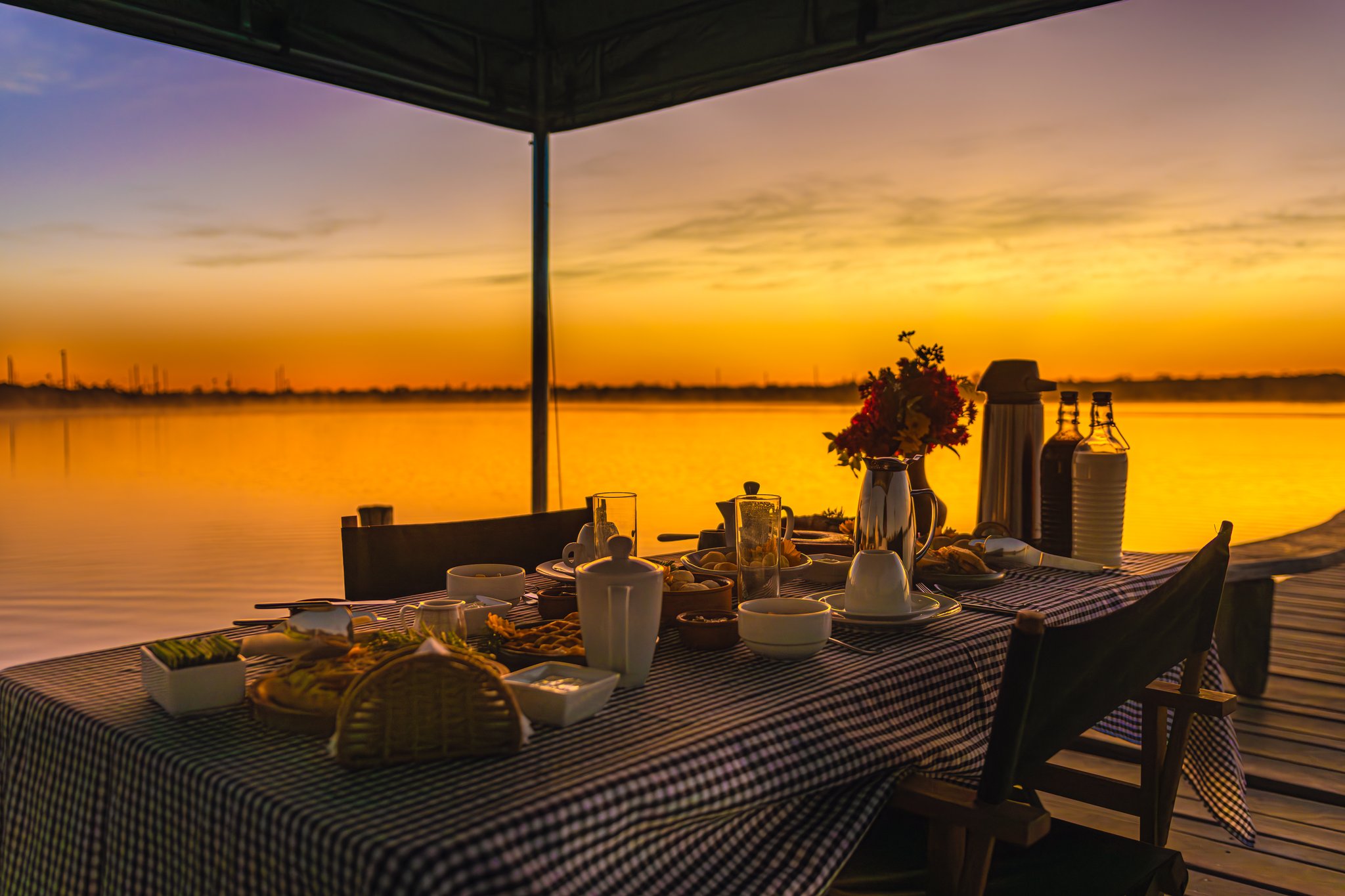 Golden hour at the pier — breakfast with a view worth waking up for. Photo: Albori Ribeiro A fully set breakfast table on a wooden pier at Pousada Trijunção, with a gingham tablecloth, fresh flowers, coffee pots and fruit, silhouetted against a vivid golden sunrise over a glassy lake