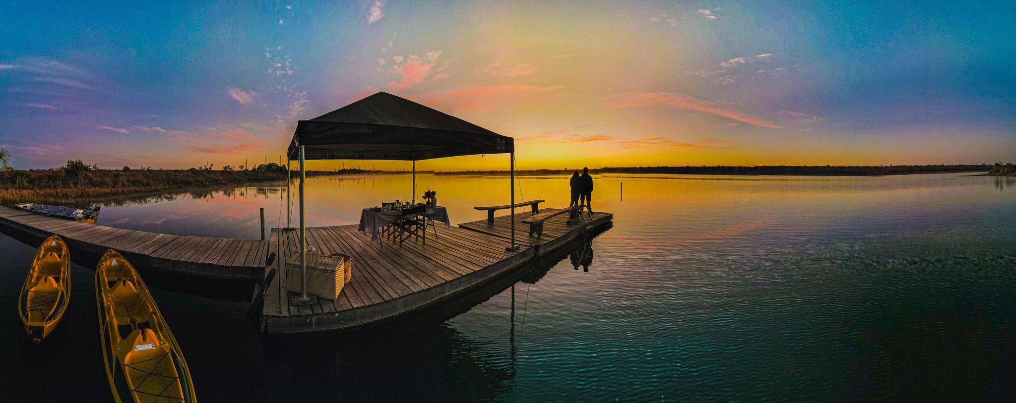 Morning light on the water — breakfast at the pier before the Cerrado wakes up. A couple silhouetted at the end of a wooden pier at Pousada Trijunção, a canopied breakfast table behind them and kayaks moored to the left, as a golden sunrise blazes across the glassy lake
