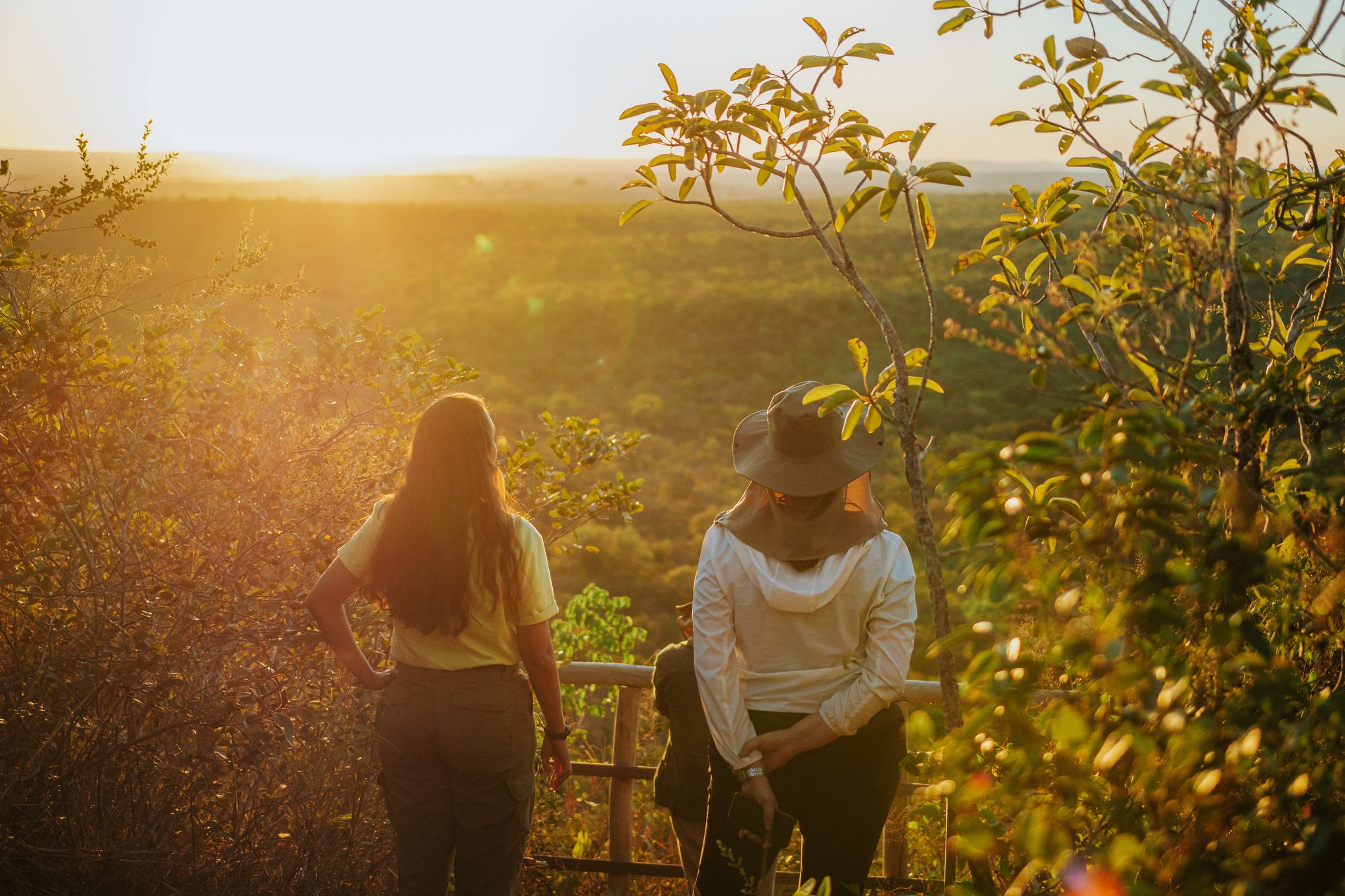 Golden hour over the Cerrado — a moment worth every mile. Photo: Albori Ribeiro Two women standing at a wooden railing viewpoint at Pousada Trijunção, gazing over a vast golden-lit Cerrado landscape at sunset