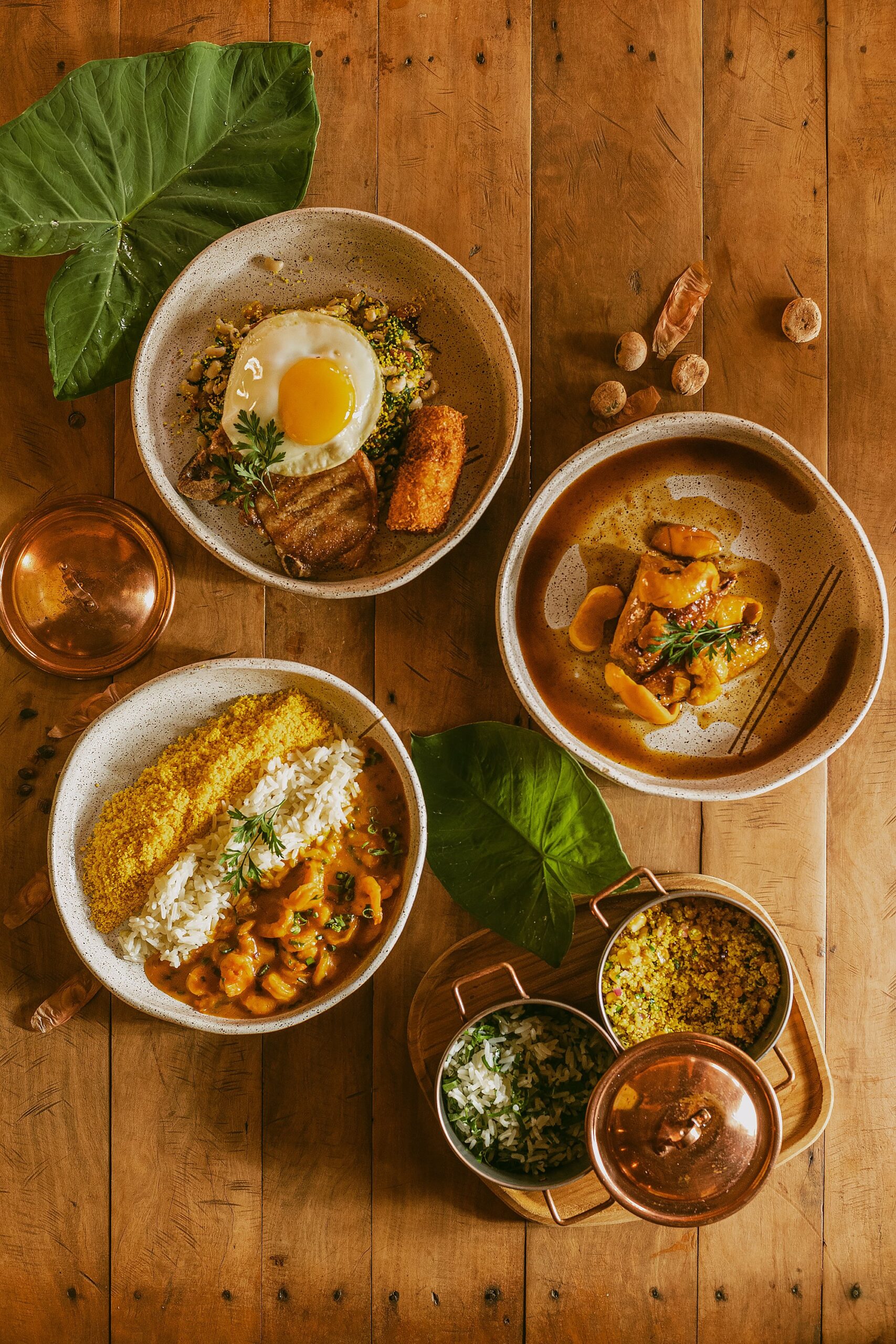 Farm-to-table flavours rooted in the Cerrado — every meal at Trijunção tells a story. Four ceramic bowls of traditional Brazilian dishes served at Pousada Trijunção, including shrimp moqueca with rice and farofa, a fried-egg rice bowl with crispy pork belly, a rich meat stew, and copper pots of sides on a warm wooden table