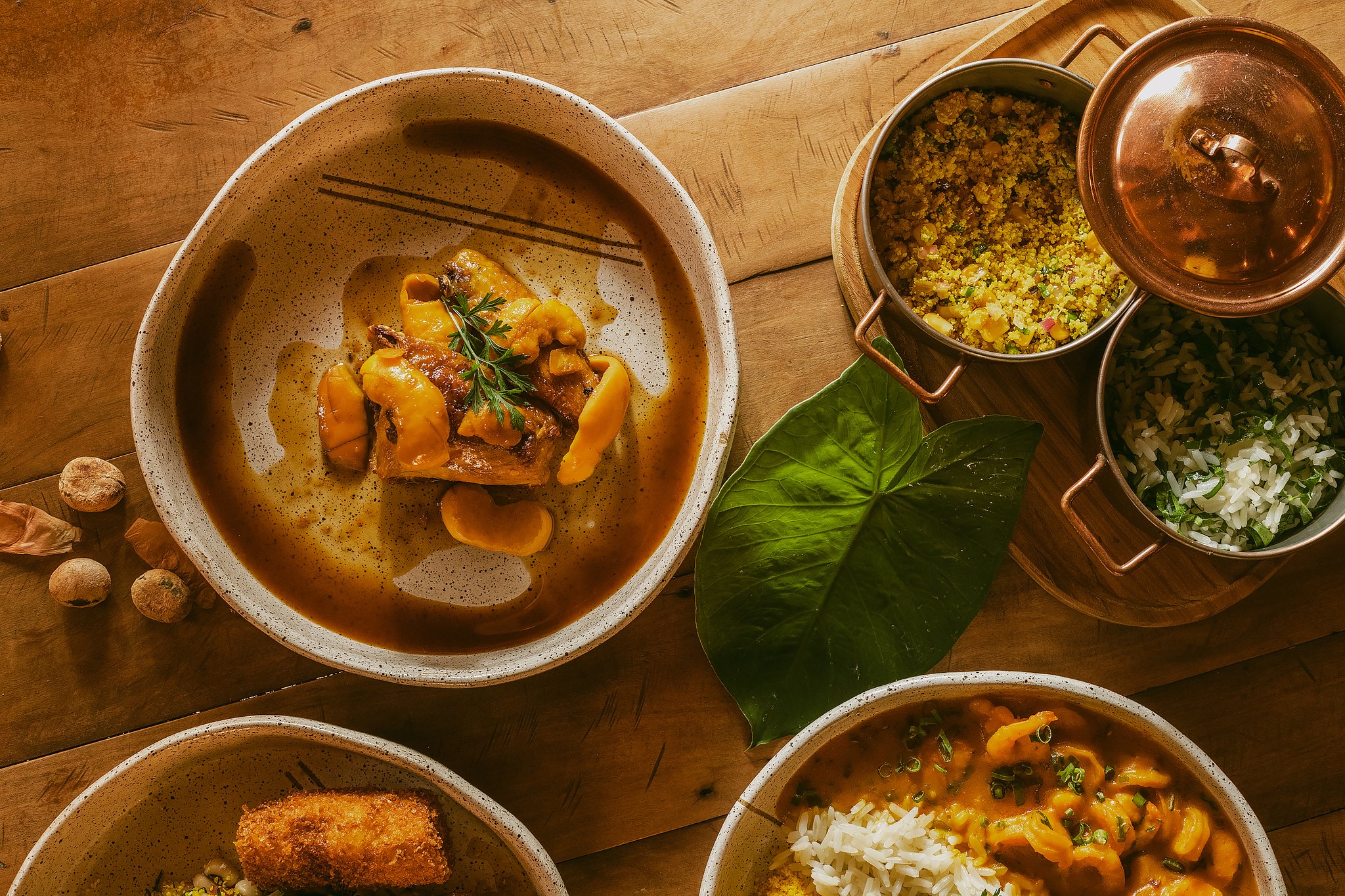 Farm-fresh flavours of the Cerrado, served with warmth and soul. Overhead view of a Pousada Trijunção meal spread featuring a rich braised meat stew in a speckled ceramic bowl, golden farofa and herb rice in copper pots, and a creamy seafood stew with white rice, all arranged on a rustic wooden table with a tropical leaf