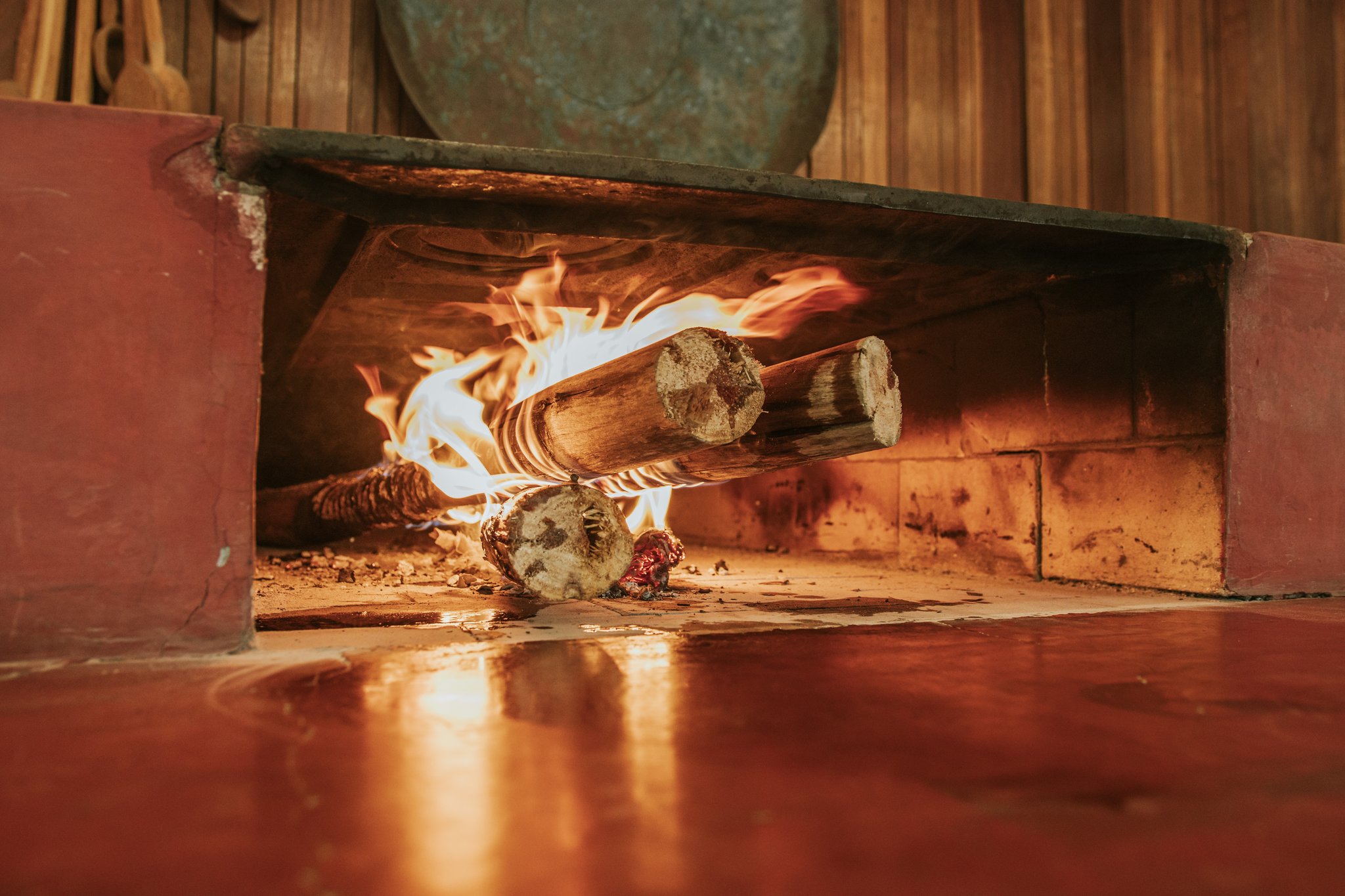 Slow fire, real flavour — the heart of the kitchen at Trijunção. Photo: Albori Ribeiro Thick wooden logs burning with open flames inside a traditional wood-fired stone hearth at Pousada Trijunção, with warm firelight reflecting off the polished red floor
