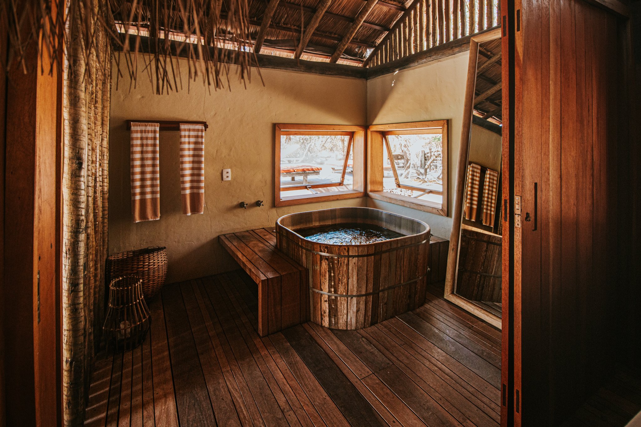 Soak in a handcrafted barrel tub while the Cerrado unfolds just beyond your window. Photo: Albori Ribeiro Rustic wooden barrel bathtub inside a Pousada Trijunção suite with thatched ceiling, warm timber floors and open windows overlooking the Cerrado landscape