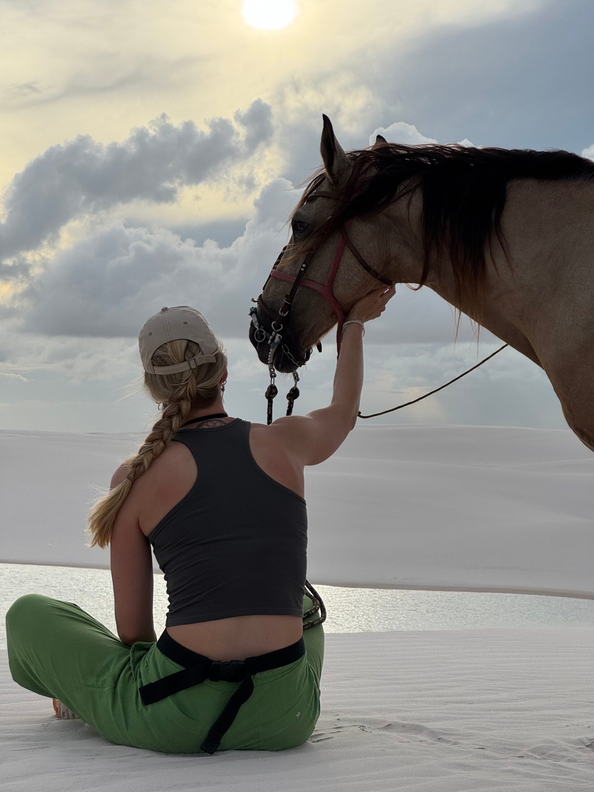 A rider in a cap pauses to stroke her horse's nose on the white dunes of Lençóis Maranhenses at sunset.