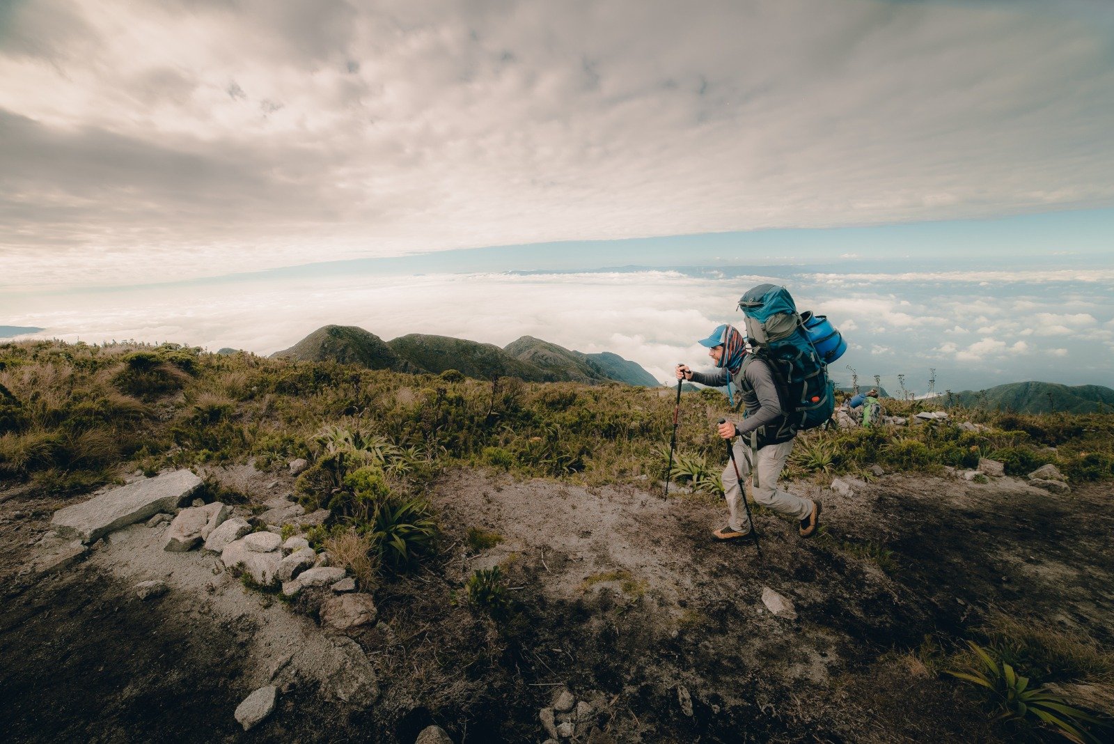 A hiker with a full backpack and trekking poles climbs a rocky high-altitude ridge on the Serra Fina Trekking route, with a sweeping sea of clouds and Mantiqueira peaks stretching to the horizon