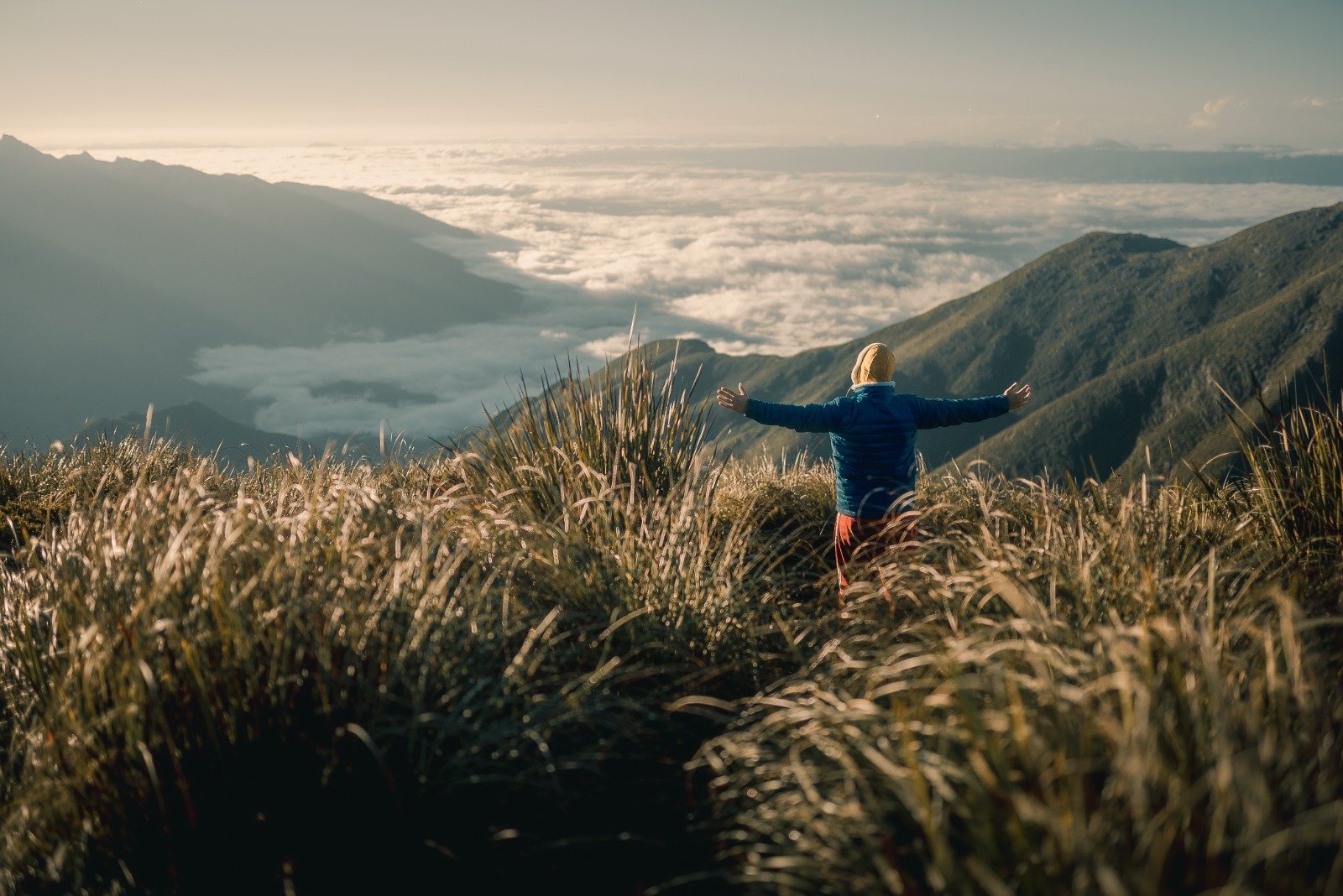 A hiker in a blue jacket stands arms wide open on a Serra Fina Trekking ridge, surrounded by golden grass with a sea of clouds filling the Mantiqueira valleys below