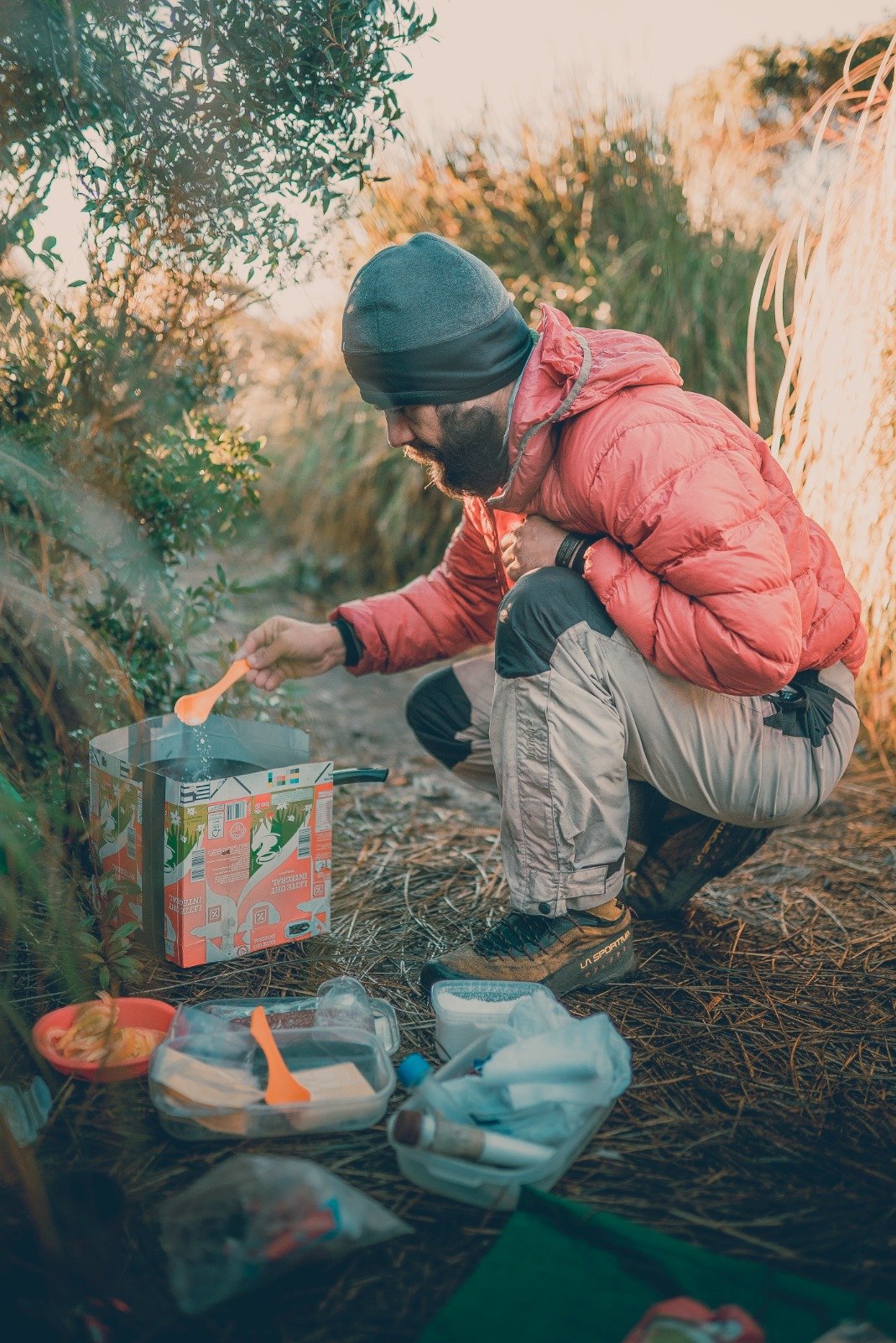 A bearded trekker in a red down jacket crouches over a portable camp stove on a dry grass clearing in the Serra da Mantiqueira, stirring a meal with an orange spoon during Serra Fina Trekking