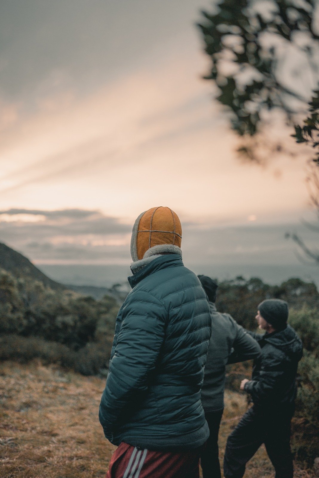 First light over the Mantiqueira — worth every cold, early morning. Two hikers in heavy winter layers and a sheepskin hat stand on an exposed ridge during Serra Fina Trekking, gazing over misty mountain valleys beneath a soft pastel sunrise sky in the Serra da Mantiqueira