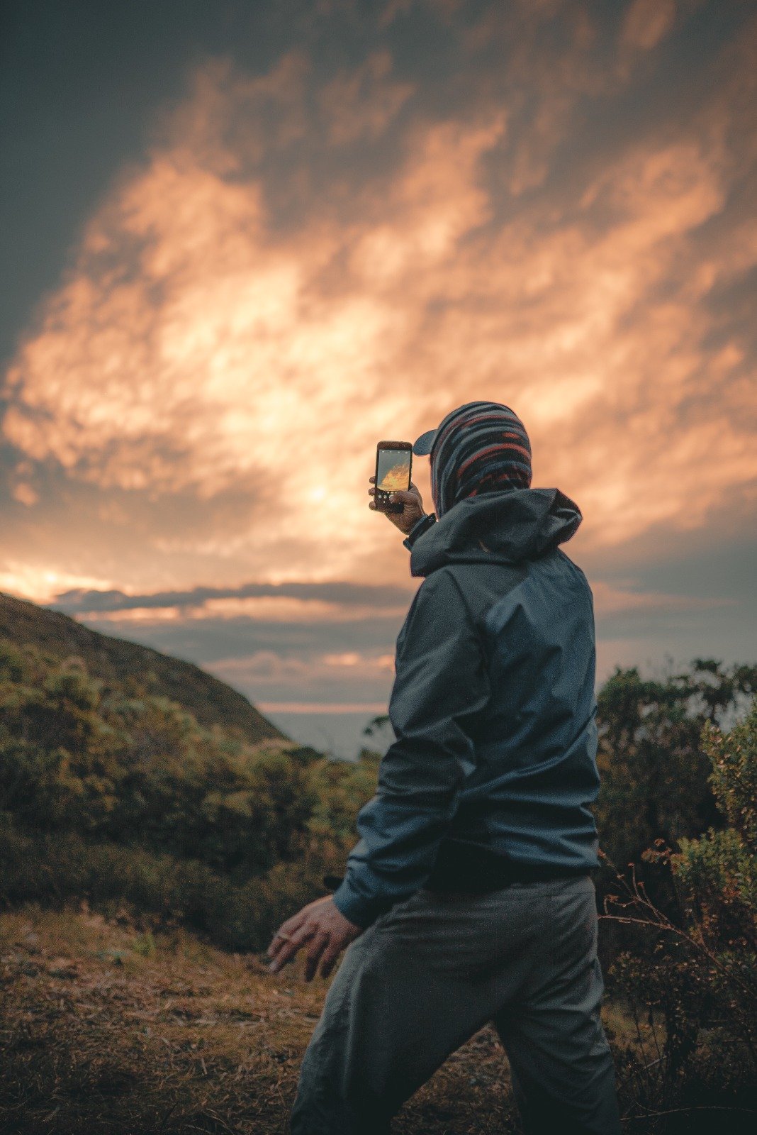 Chasing golden skies above the Mantiqueira — no filter needed. A hiker in a jacket and striped beanie photographs a dramatic orange and gold sunset sky above the Serra da Mantiqueira ridgeline during a Serra Fina Trekking expedition