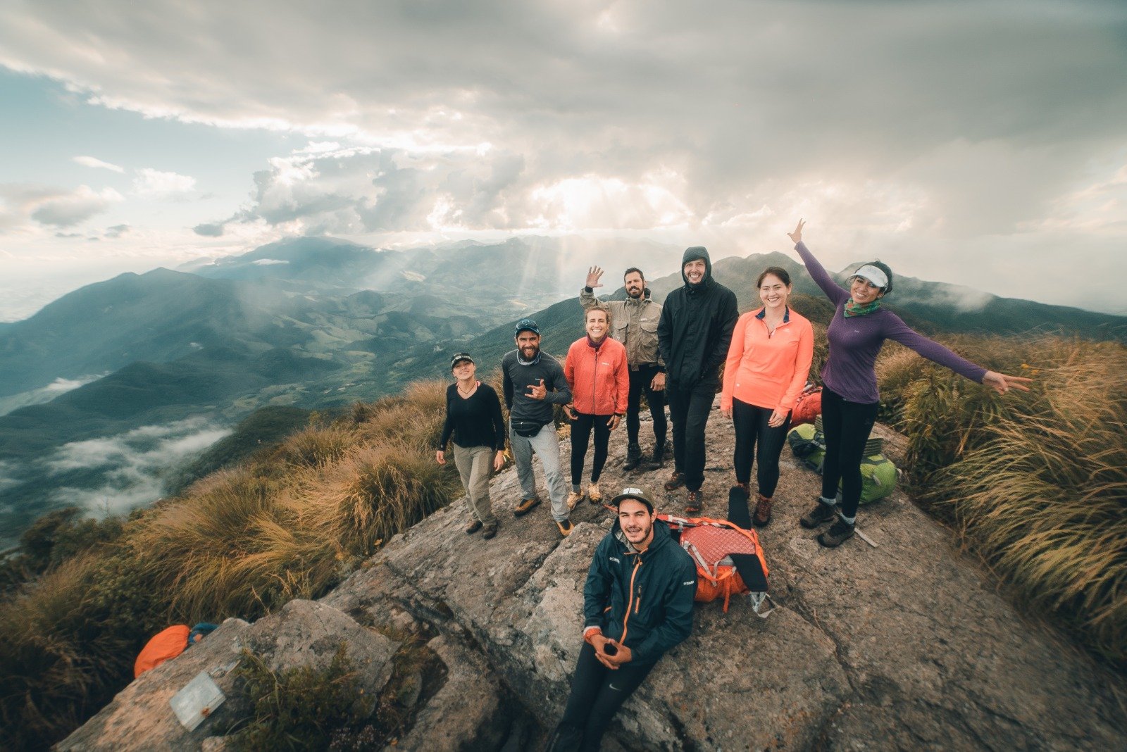 Summit smiles, open skies — the Mantiqueira at your feet. Eight hikers celebrate on a rocky summit during the Serra Fina Trekking, with dramatic cloud-filled skies and the rolling ridges of the Serra da Mantiqueira stretching to the horizon