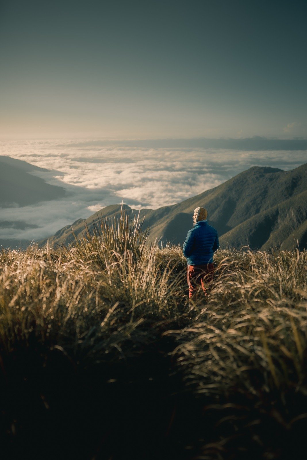 Above the clouds, below the sky — the Serra da Mantiqueira at its most humbling. A hiker in a blue down jacket stands among golden highland grasses on the Serra Fina Trekking route, gazing over a vast sea of clouds filling the valleys of the Serra da Mantiqueira