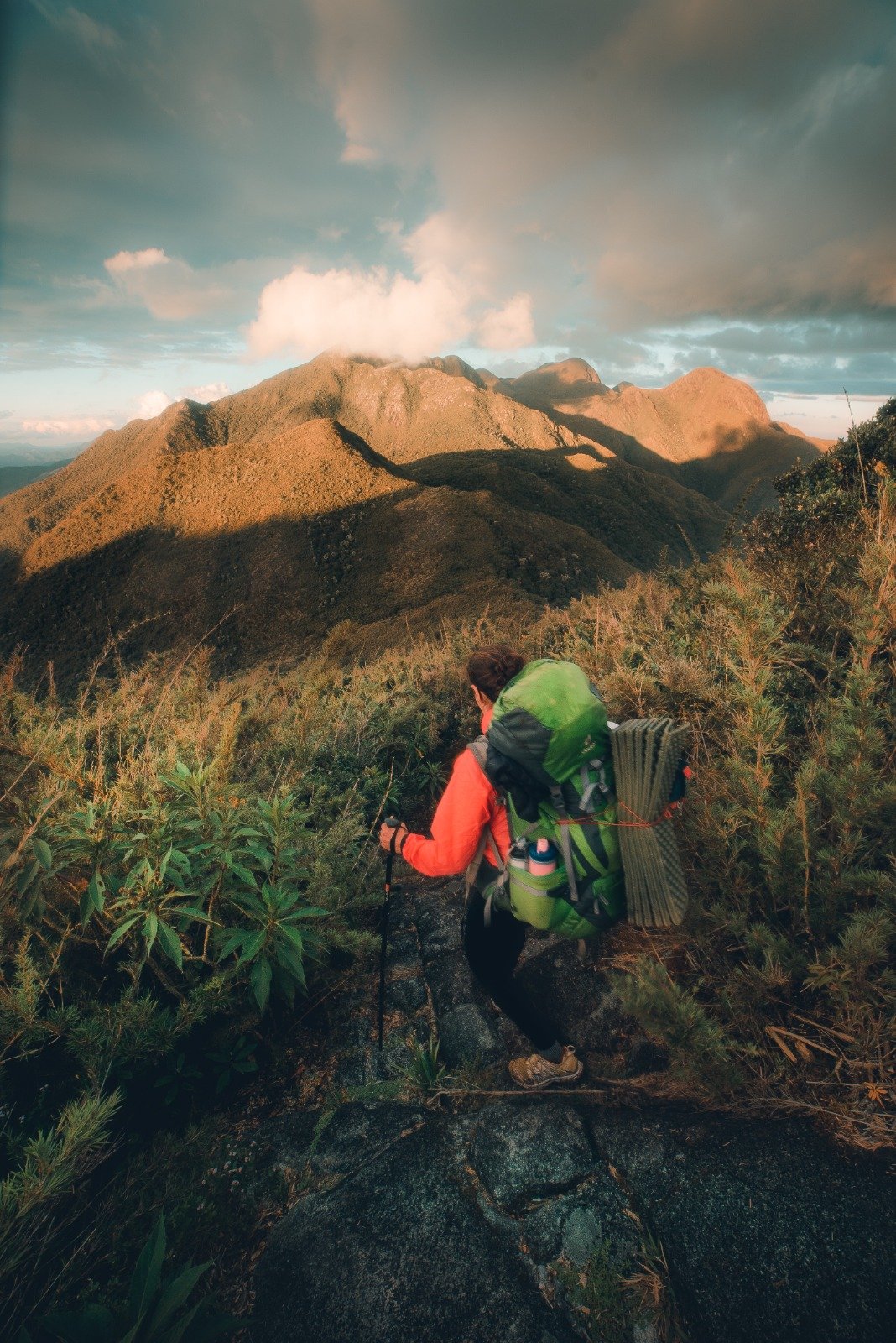 The ridge opens up — and the Mantiqueira takes your breath away. A backpacker in an orange jacket carrying a full pack pauses on a rocky ridge during Serra Fina Trekking, with golden-lit mountain summits of the Serra da Mantiqueira stretching under a dramatic cloudy sky