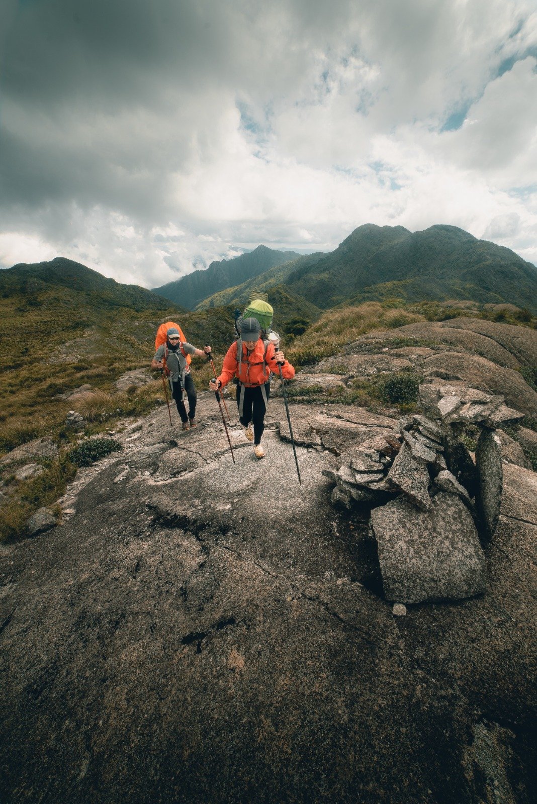 Every step earned on the high ridges of Serra Fina. Two backpackers in orange jackets using trekking poles to navigate a granite rock slab on the Serra Fina Trekking route, with green mountain ridges and dramatic clouds filling the Serra da Mantiqueira skyline behind them