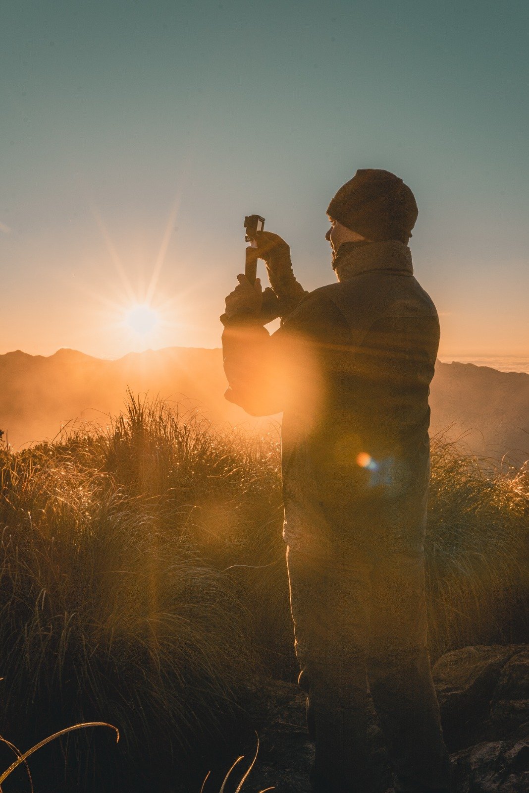 First light on the Mantiqueira — a moment worth every steep metre. A hiker in a beanie and puffer jacket photographs the golden sunrise over misty Serra da Mantiqueira ridges during a Serra Fina trekking expedition, wild grasses glowing in the foreground