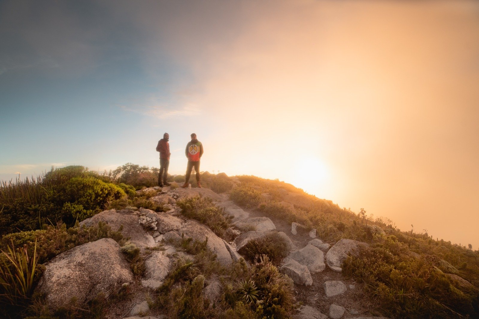 Standing at the top — where the Serra da Mantiqueira glows gold at dawn. Two hikers silhouetted against a glowing golden sunrise on a rocky Serra Fina Trekking summit, surrounded by high-altitude Atlantic forest vegetation in the Serra da Mantiqueira.