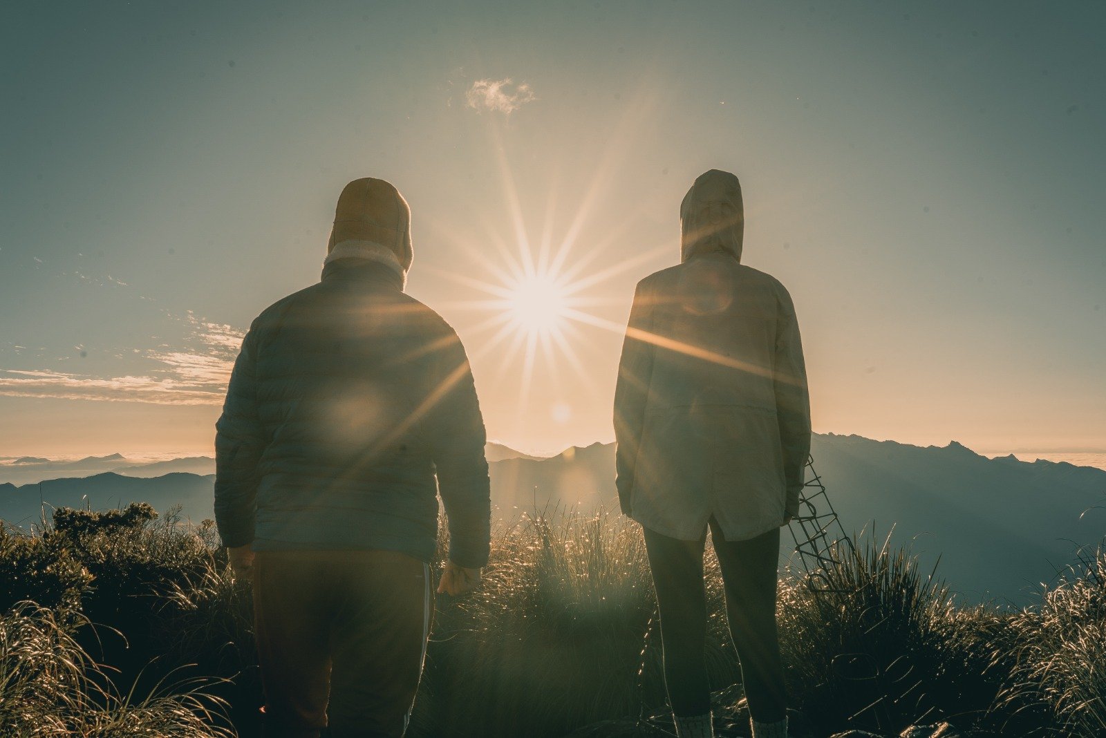 First light from the top of the world — Serra da Mantiqueira at its most raw. Two hooded hikers silhouetted against a brilliant starburst sunrise on a Serra Fina Trekking summit, with layers of Mantiqueira mountain ridges stretching to the horizon