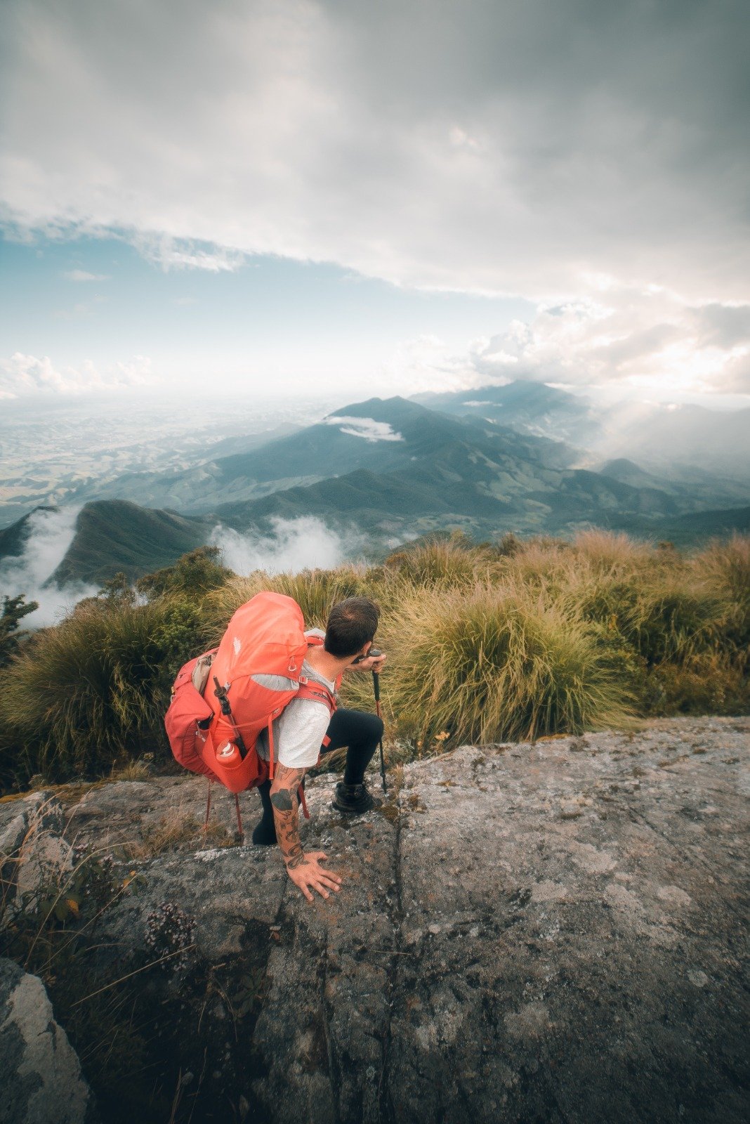 On top of the world — clouds, ridges and silence all the way to the horizon. A hiker with a large red backpack and trekking pole crouches on a rocky ledge during Serra Fina Trekking, gazing over a vast misty mountain landscape in the Serra da Mantiqueira