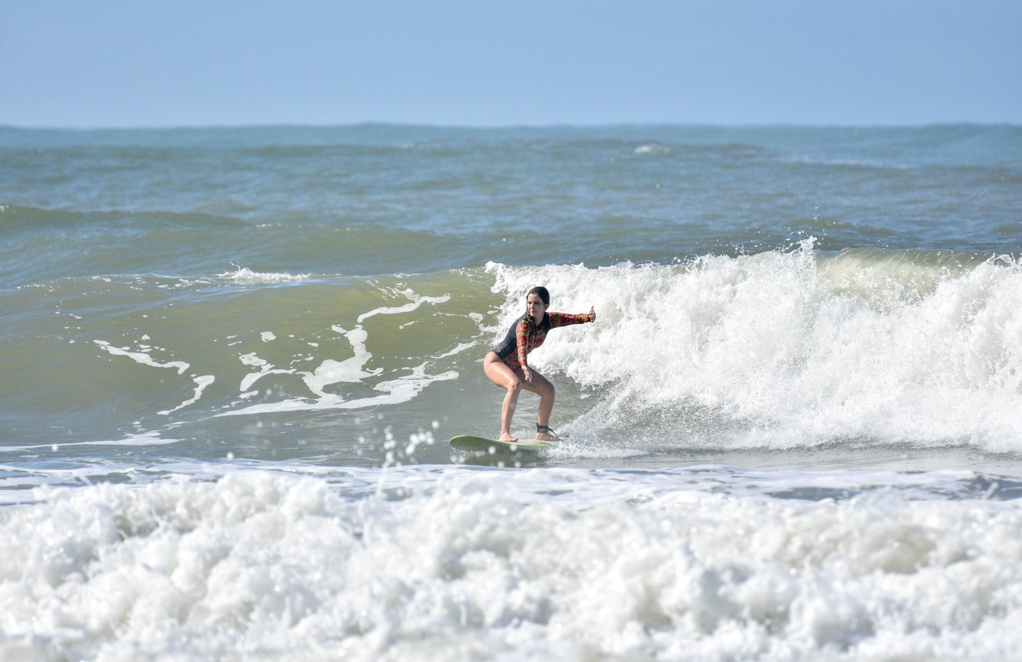 A woman crouches low on a surfboard while taking surf lessons in Itacaré, riding a foaming wave under a clear blue sky