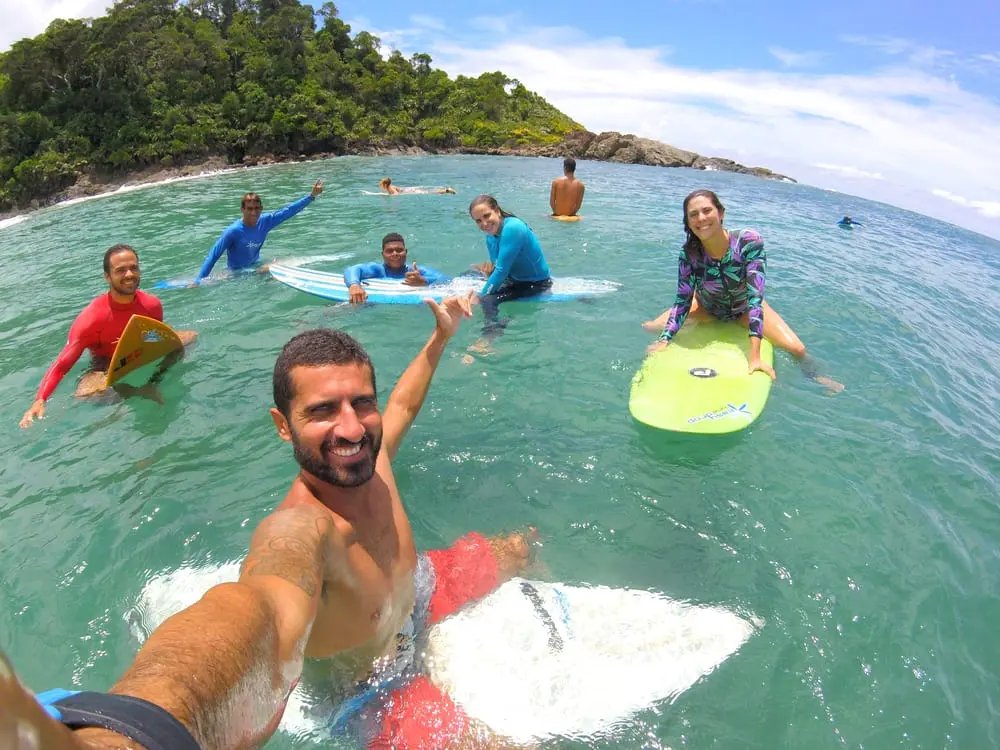 A smiling group of students and instructors floating on surfboards in the clear turquoise waters of Itacaré at this surf school in Itacaré, with a lush forested headland in the background