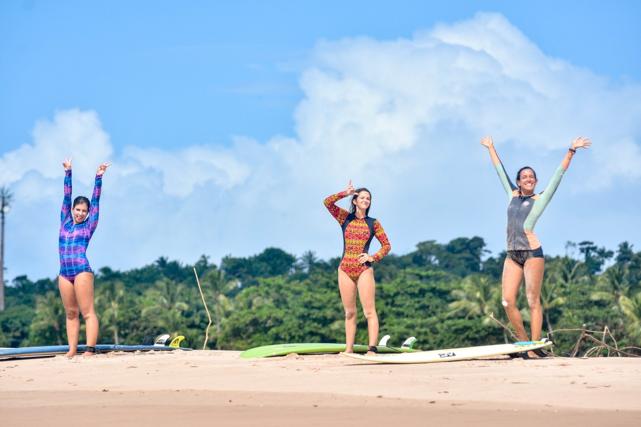 Pure stoke on the sand — the Hawaii of Brazil delivers every time. Photo: Picasa Three smiling women in colourful rash guards raise their arms in celebration on a sandy beach at this surf school in Itacaré, with surfboards on the sand and lush tropical forest under a blue sky behind them