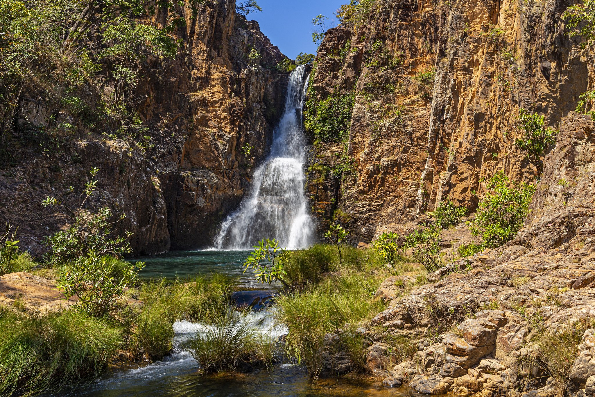 A powerful waterfall cascades down amber sandstone cliffs into a clear green pool surrounded by native Cerrado vegetation during trekking Chapada dos Veadeiros