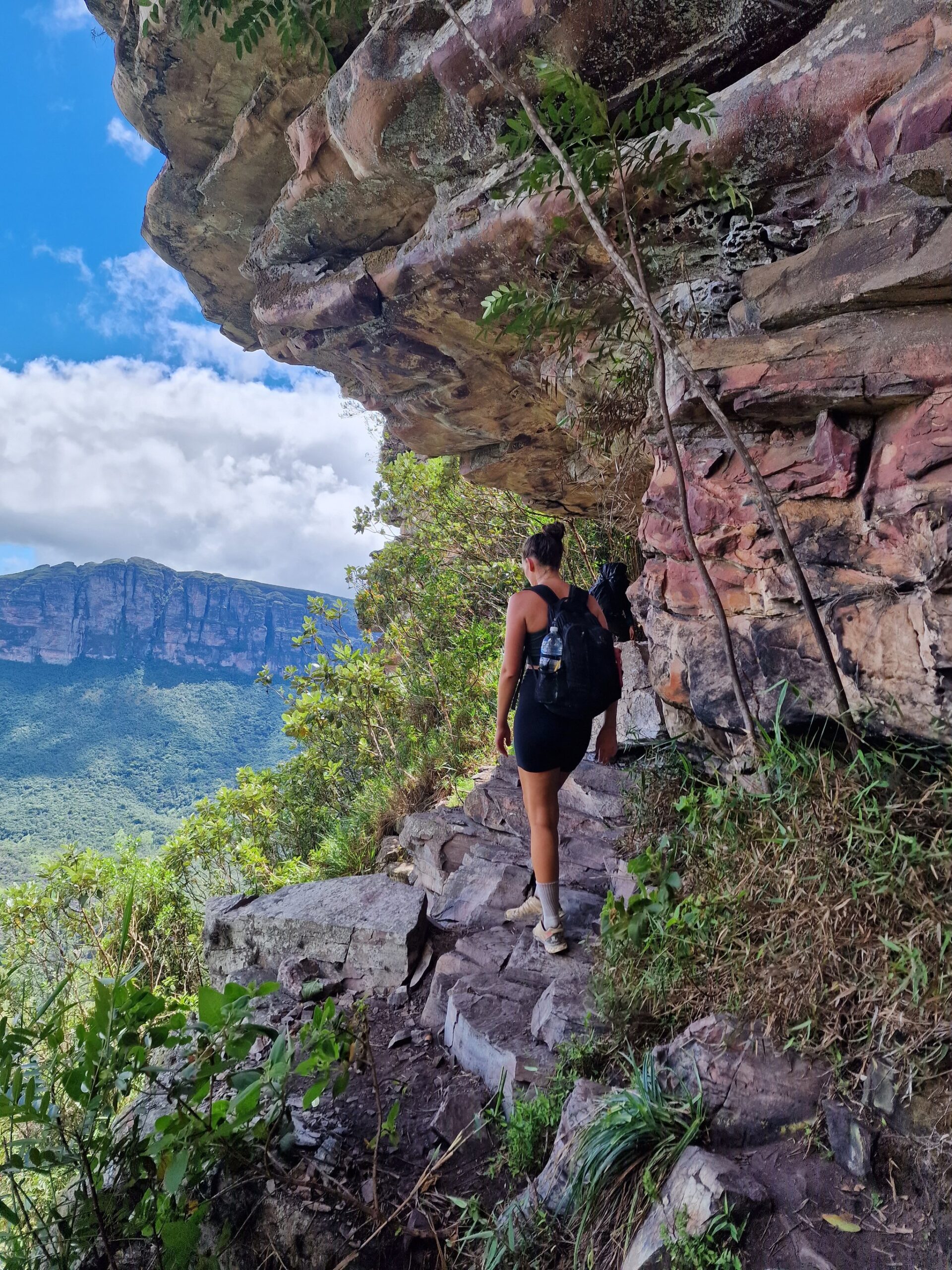 A hiker with a backpack navigates a narrow rocky ledge beneath a sandstone overhang, with a dramatic cliff-faced mountain rising above dense green forest in the background — trekking in Brazil at its most exhilarating