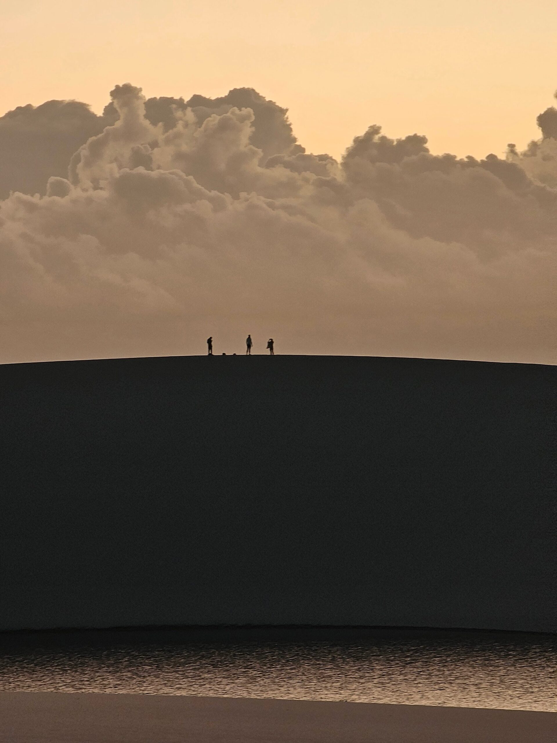 Three tiny silhouettes stand on the curved ridge of a vast sand dune at golden-hour dusk, a dramatic cumulus cloudbank rising behind them — a defining moment in trekking in Brazil's Lençóis Maranhenses.