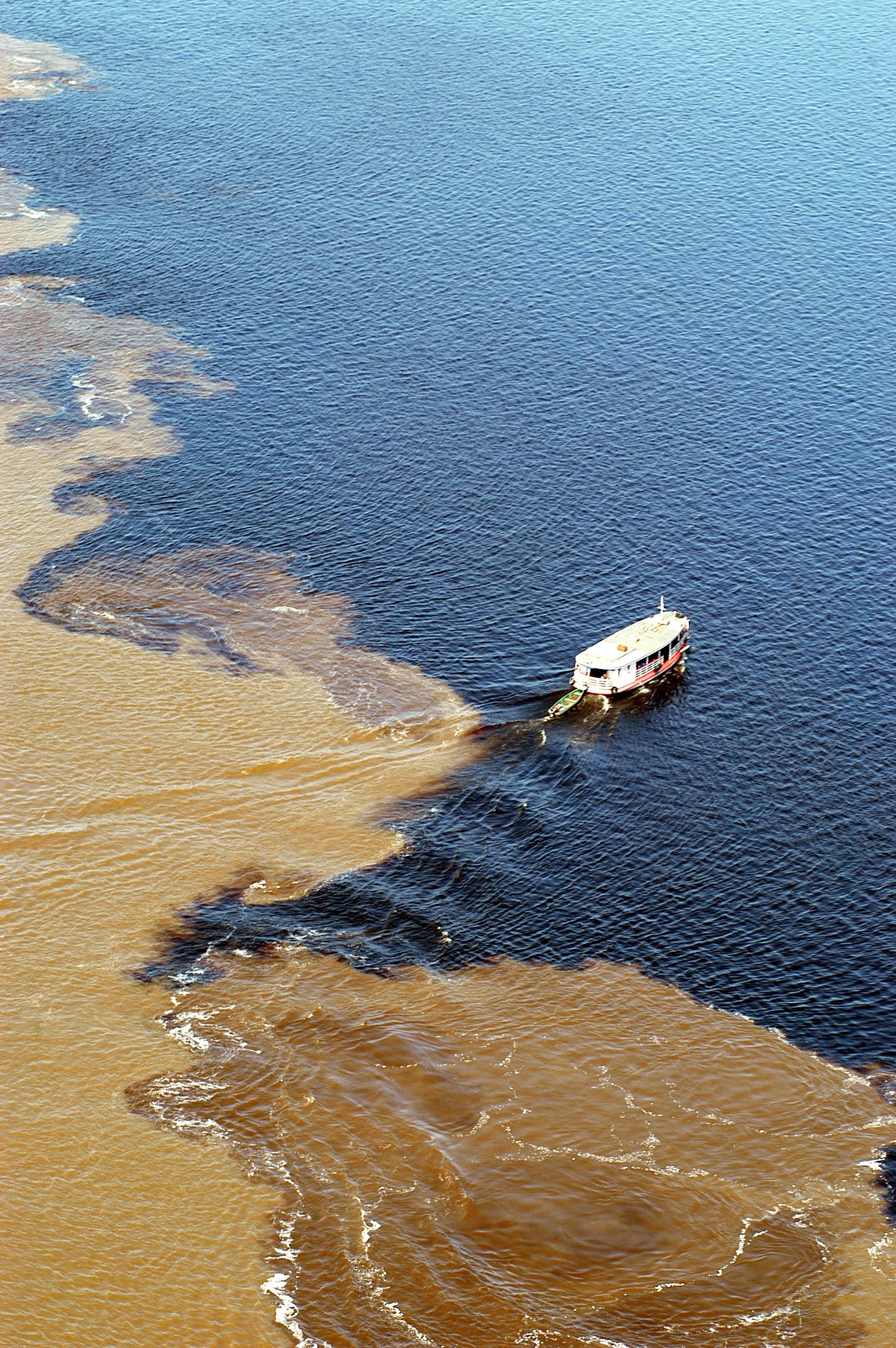 Two rivers, one boundary — the Meeting of Waters seen from above, a small vessel threading the divide. Aerial view of a river cruise vessel crossing the Meeting of Waters, where dark black-water and sandy brown-water rivers flow side by side without mixing