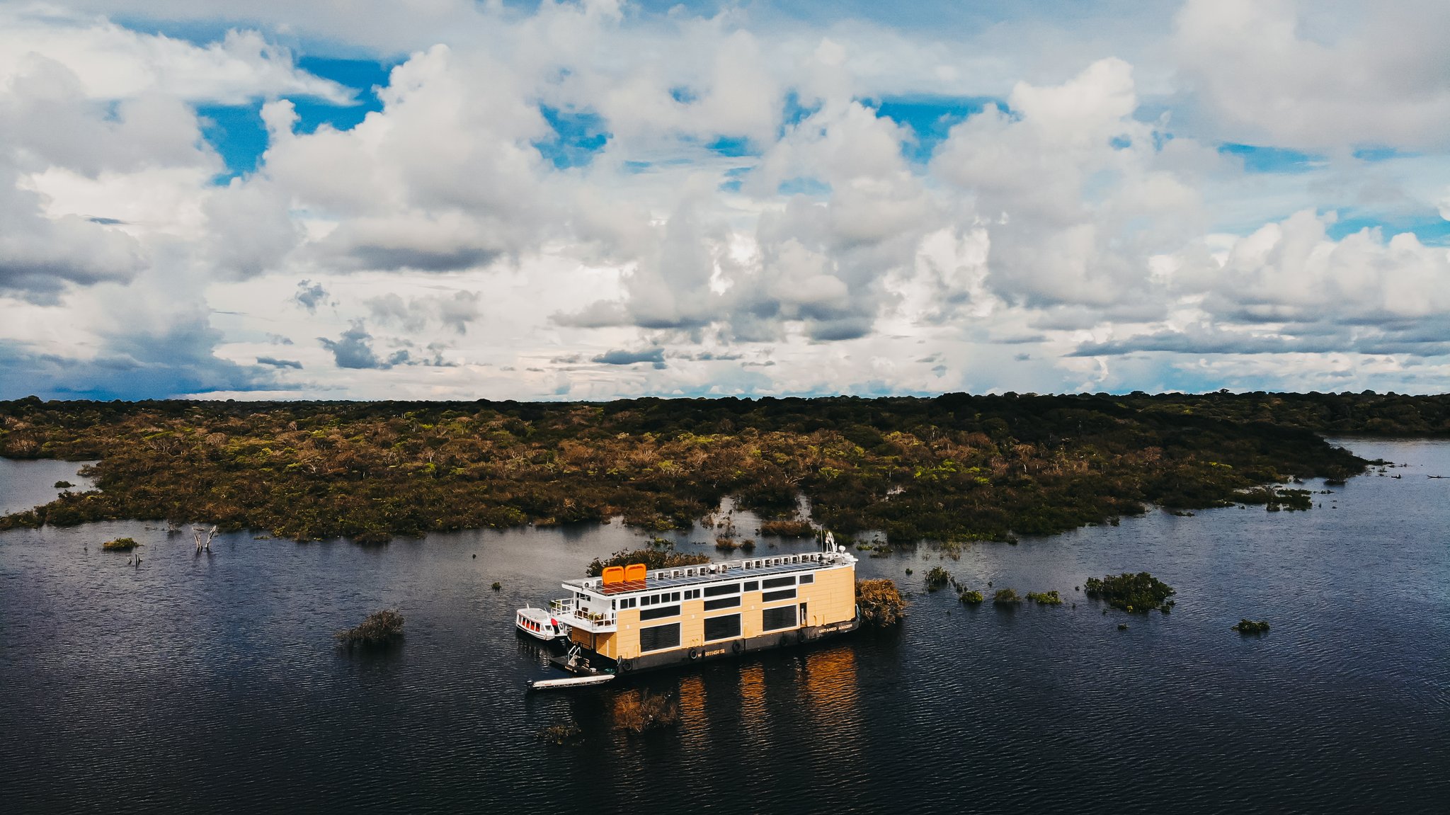 Aerial view of the untamed amazon cruise vessel moored among flooded trees and dark water beneath a dramatic cloudy sky