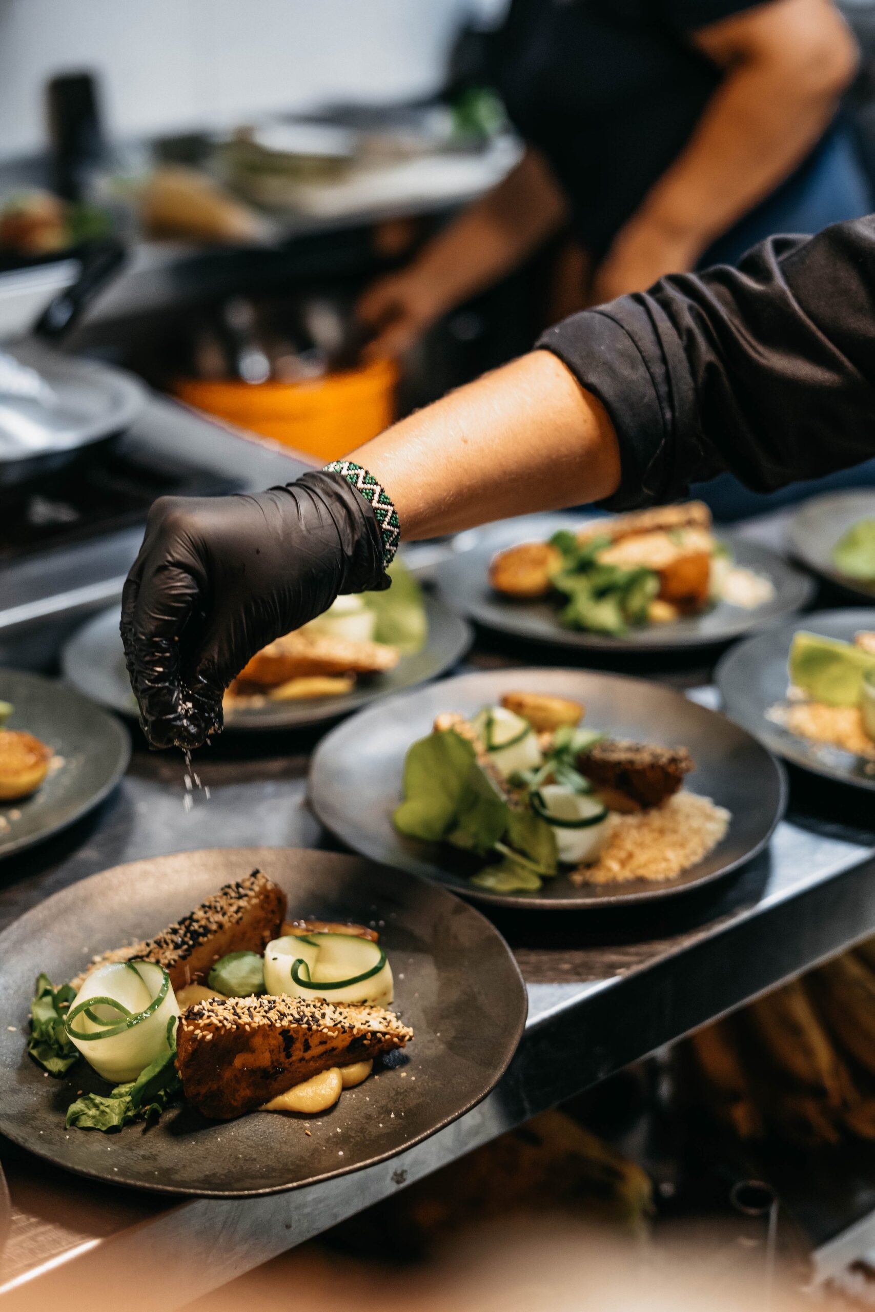 A chef wearing black gloves sprinkles seasoning over multiple elegantly plated dishes featuring sesame-crusted portions, spiralled cucumber and fresh greens, prepared aboard the untamed amazon cruise