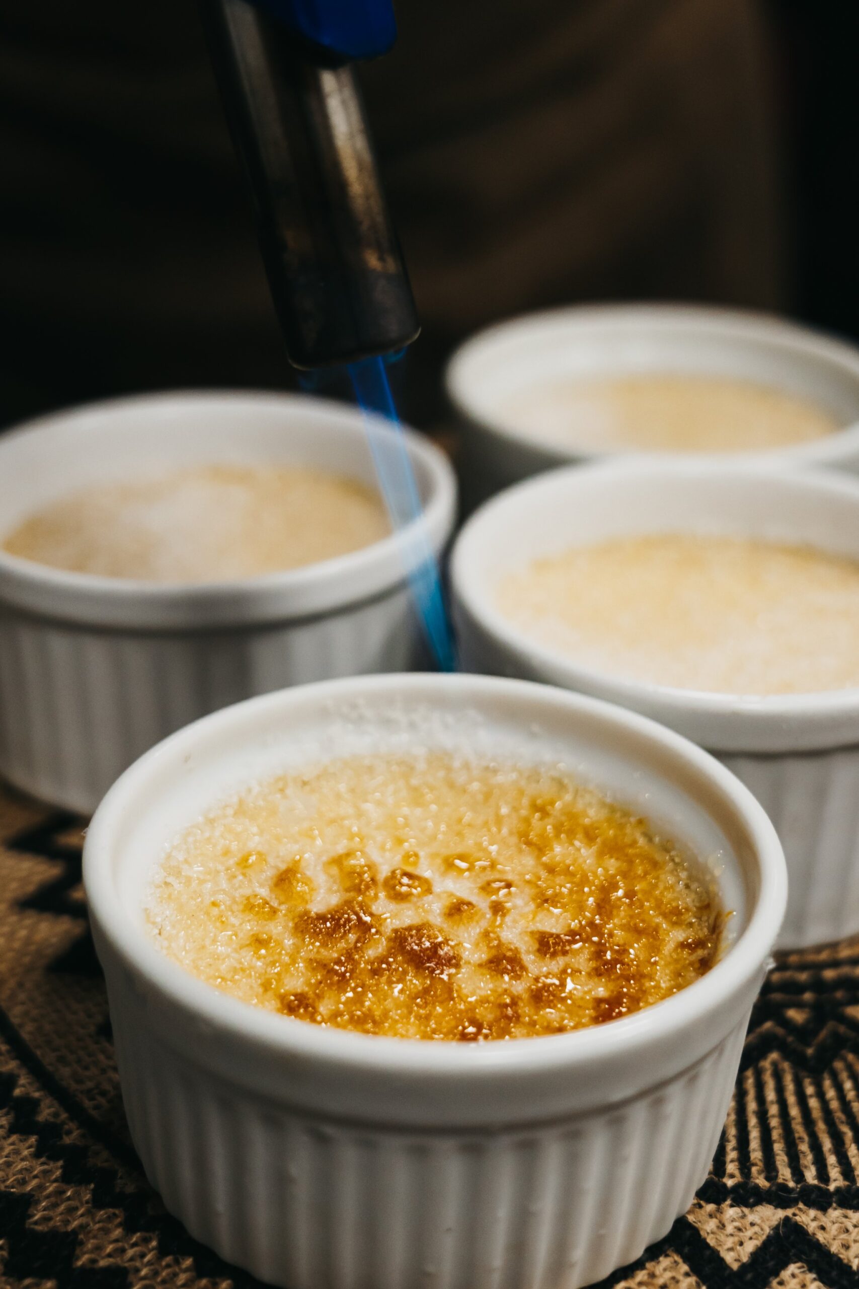 A kitchen torch caramelising the sugar crust on a crème brûlée in a white ramekin, with three more unfinished ramekins in the background, served aboard the untamed amazon cruise