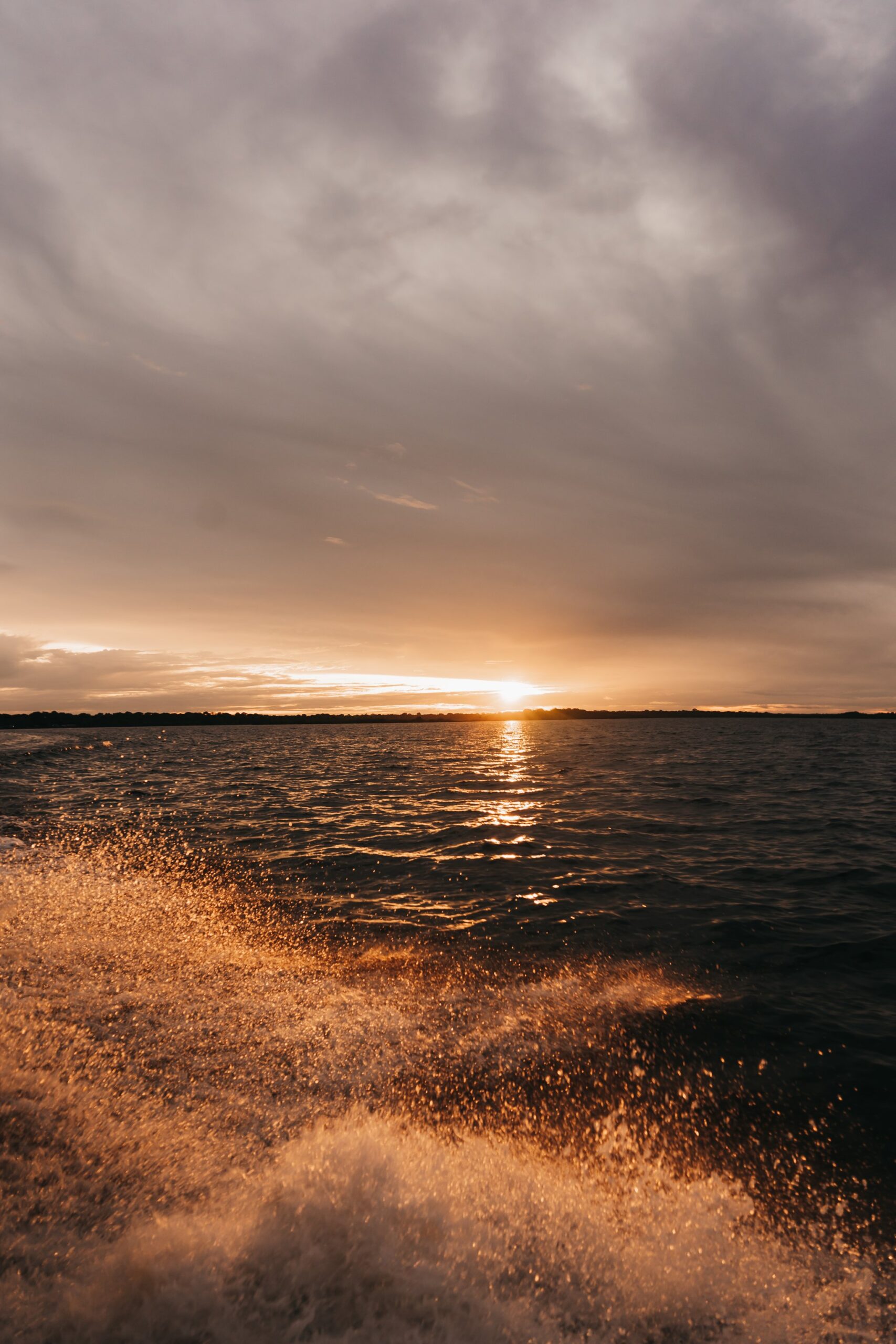 Golden boat wake glittering in the foreground as the sun sets low over a wide river on an untamed amazon cruise, with a distant treeline silhouetted beneath a dramatic cloudy sky