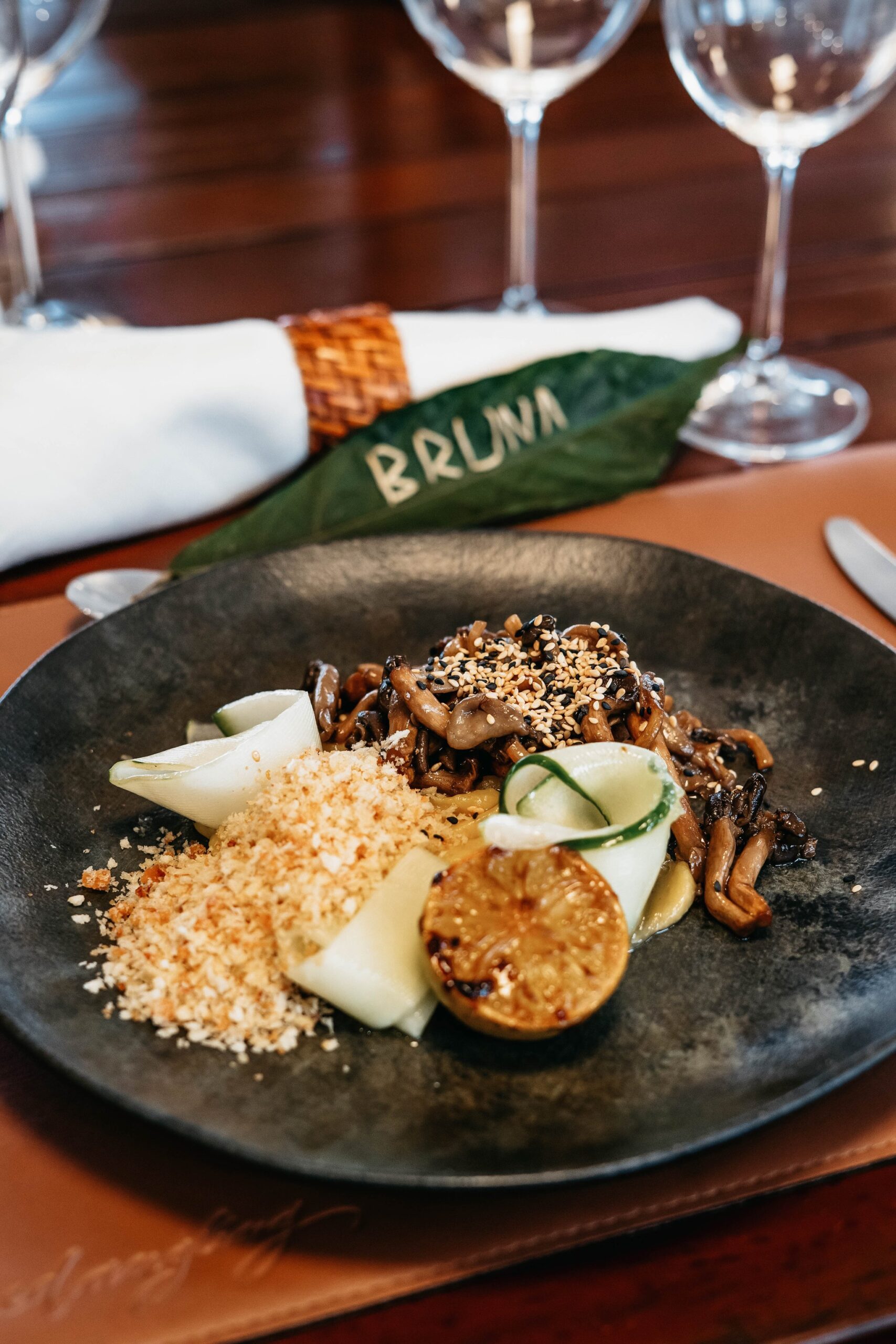 A sautéed mixed mushroom dish with sesame seeds, pickled vegetables, toasted breadcrumbs and a caramelised round vegetable slice, plated on a dark stone dish aboard the untamed amazon cruise, with a woven-ring napkin and a leaf place card reading 'Bruna' in the background