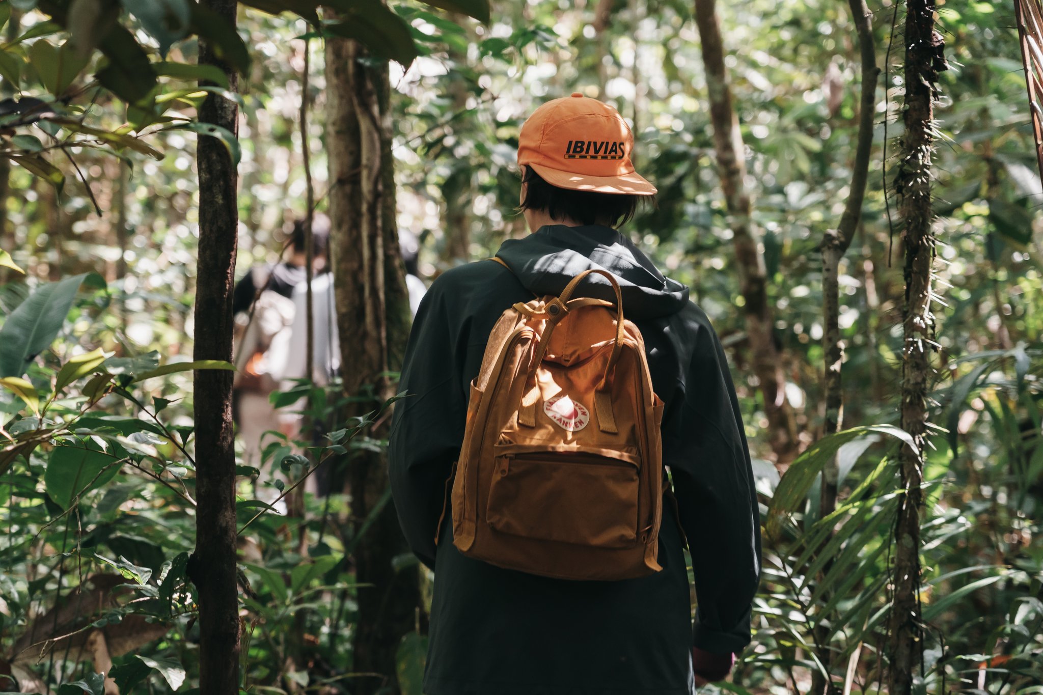 A hiker with an orange cap and tan backpack walks through dense rainforest undergrowth on an untamed amazon cruise excursion, with fellow travellers visible ahead on the trail