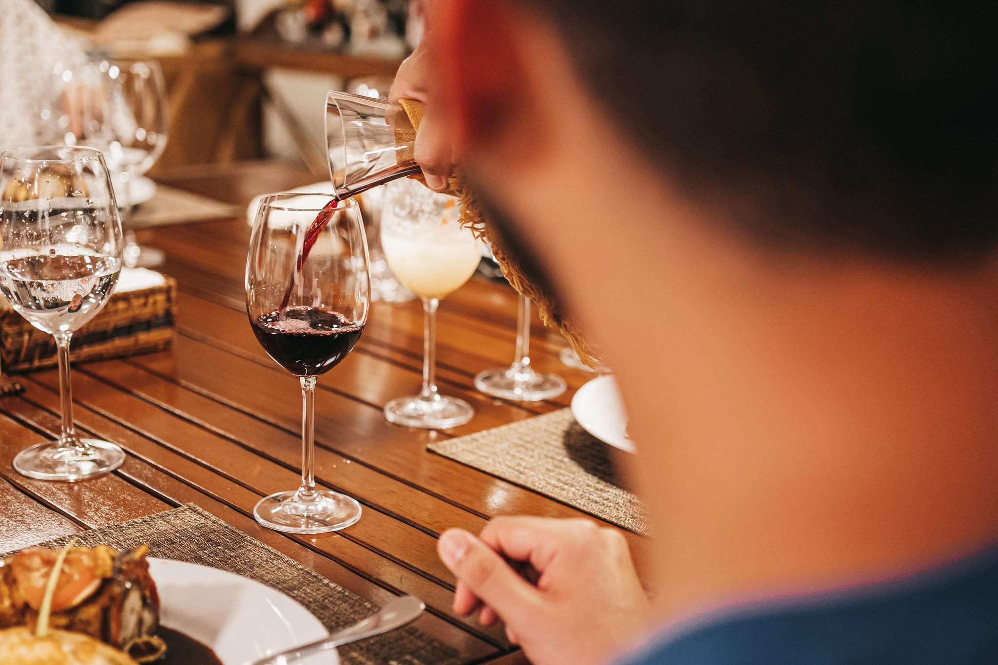Red wine being poured into a crystal glass at a elegantly set dining table aboard the untamed amazon cruise, with cocktails and plated food visible alongside
