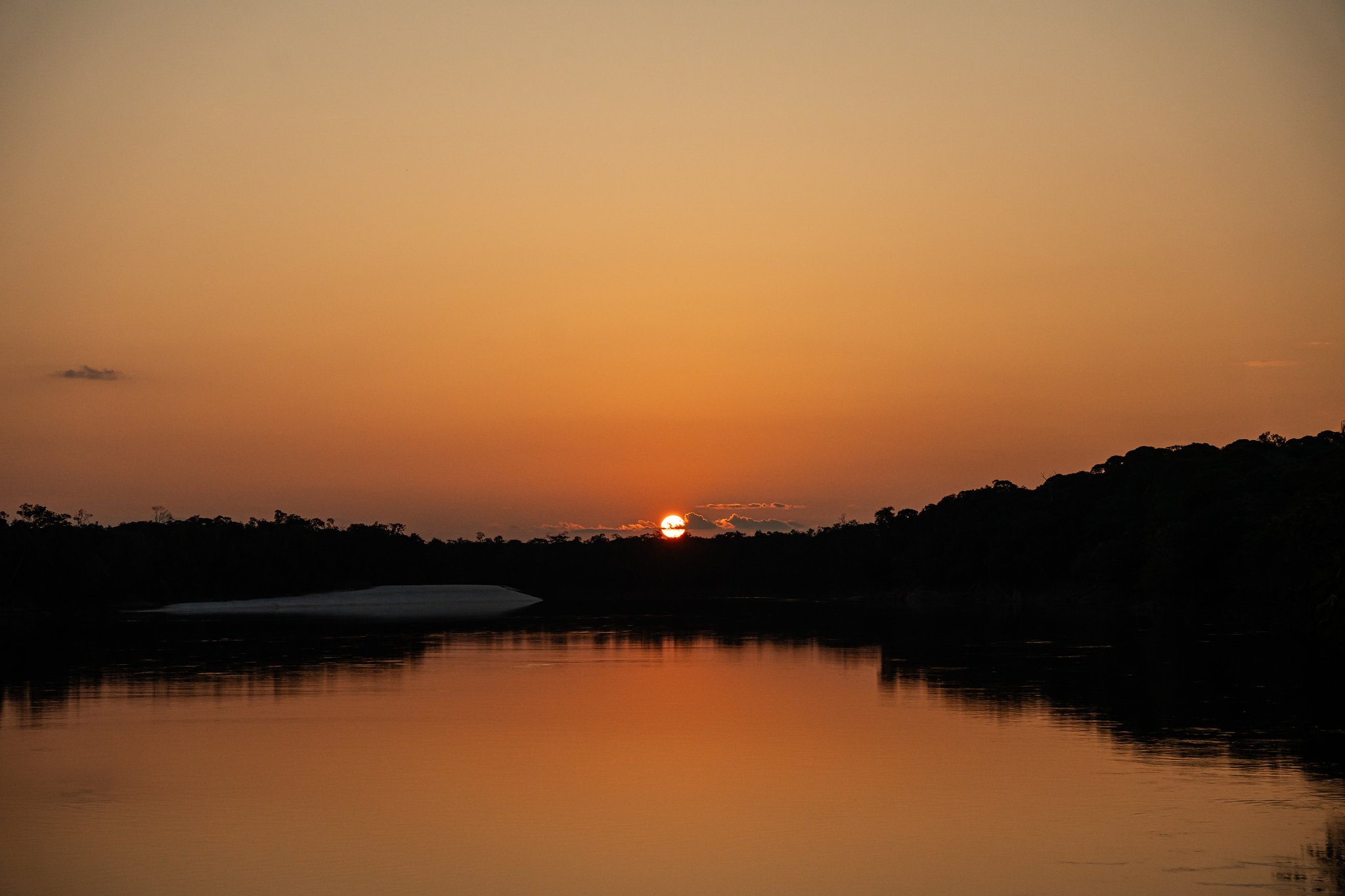 A glowing orange sun sinks behind a dark treeline silhouette on an untamed Amazon cruise, its reflection shimmering across a calm river with a white sandbar visible on the left bank