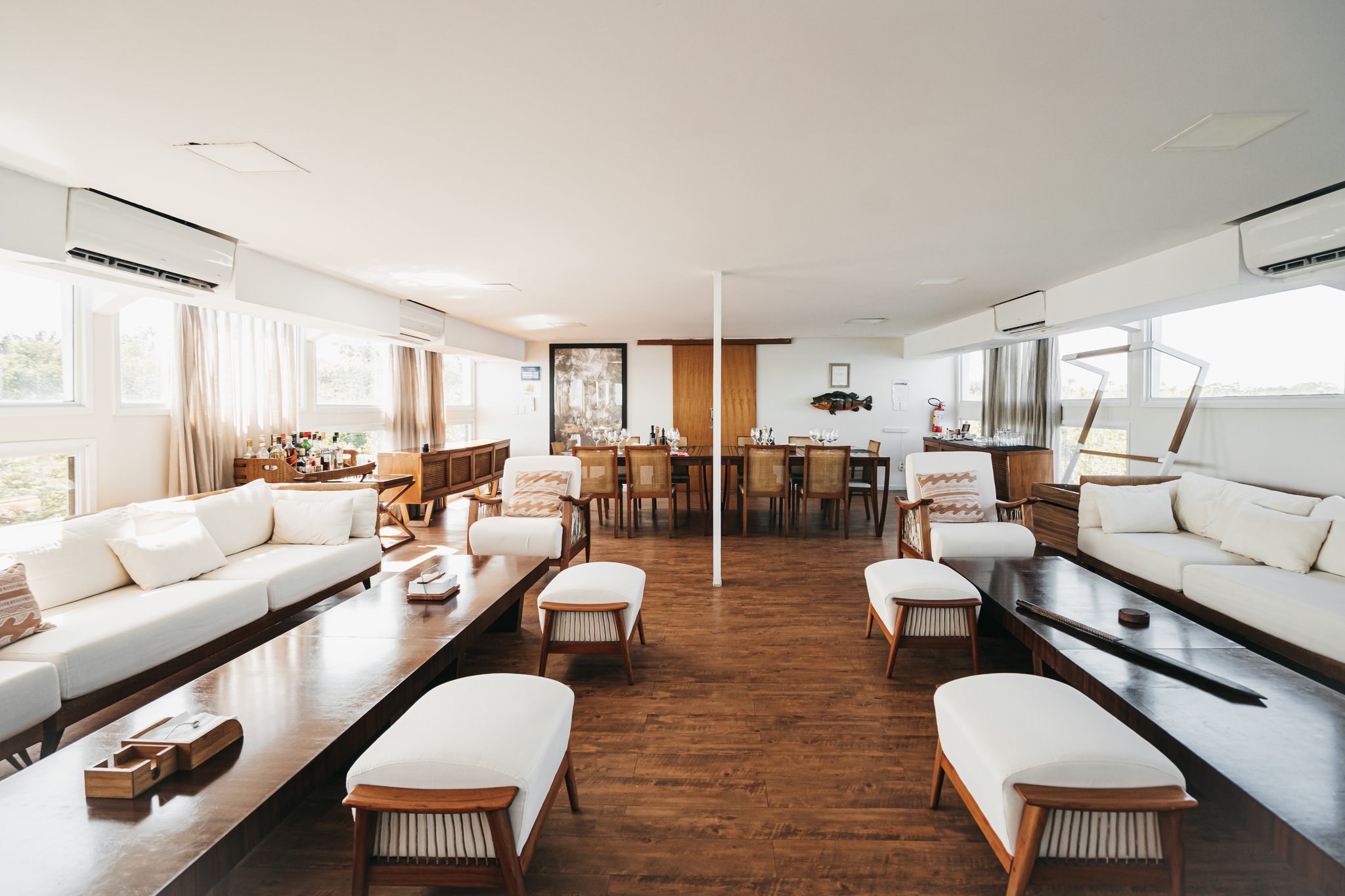 Spacious social deck of the untamed amazon cruise featuring white upholstered sofas, teak armchairs, ottomans, dark wood coffee tables, and a dining area set with glassware at the far end