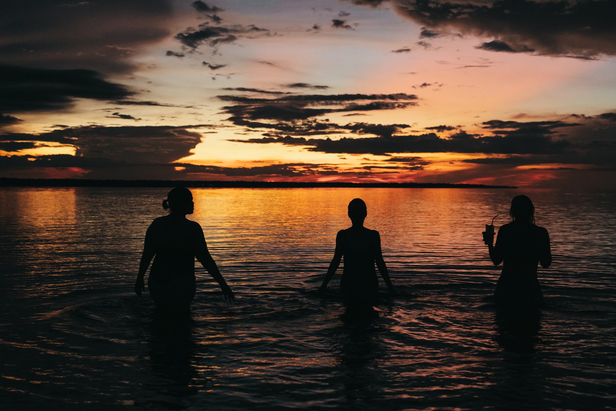 Three guests silhouetted waist-deep in calm river water during a vivid orange and red sunset on an untamed amazon cruise