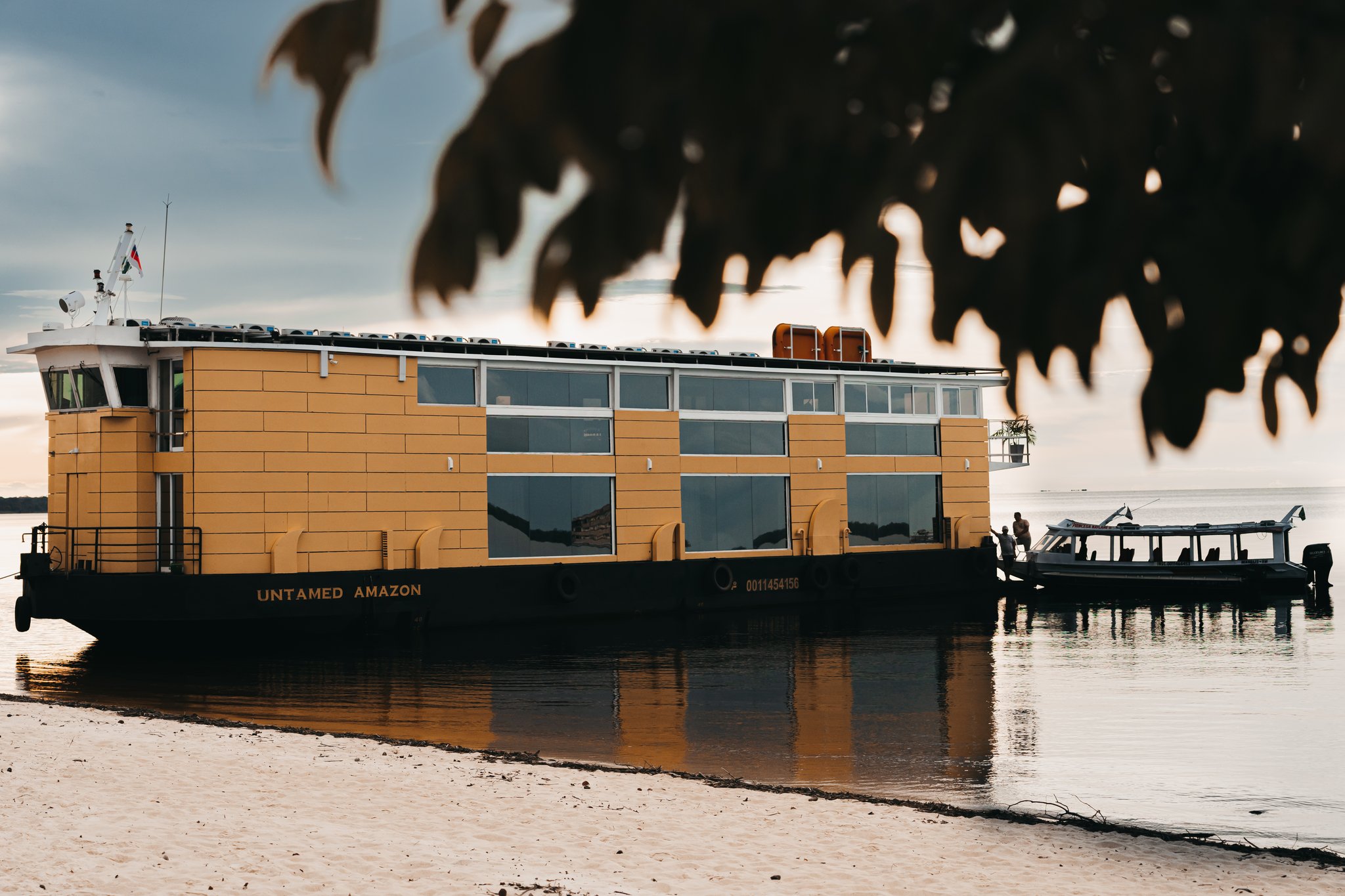 The Untamed Amazon cruise vessel moored at a sandy riverbank at sunset, its yellow hull and panoramic cabin windows reflected in the calm water, with a smaller excursion boat alongside
