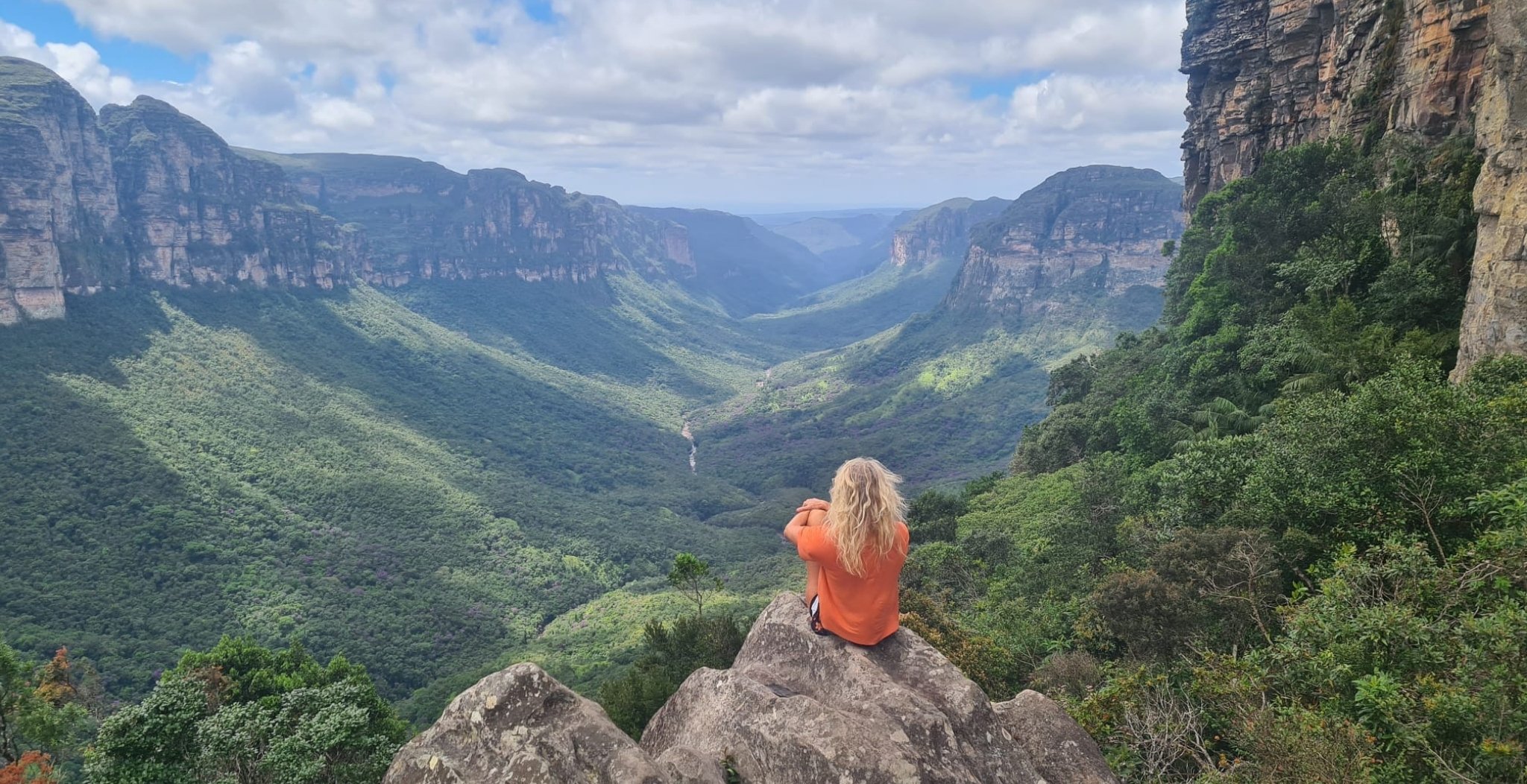 A hiker in an orange top sits on a rock outcrop overlooking the vast green canyon of the Vale do Pati Trek, with sandstone cliffs and a distant waterfall visible below