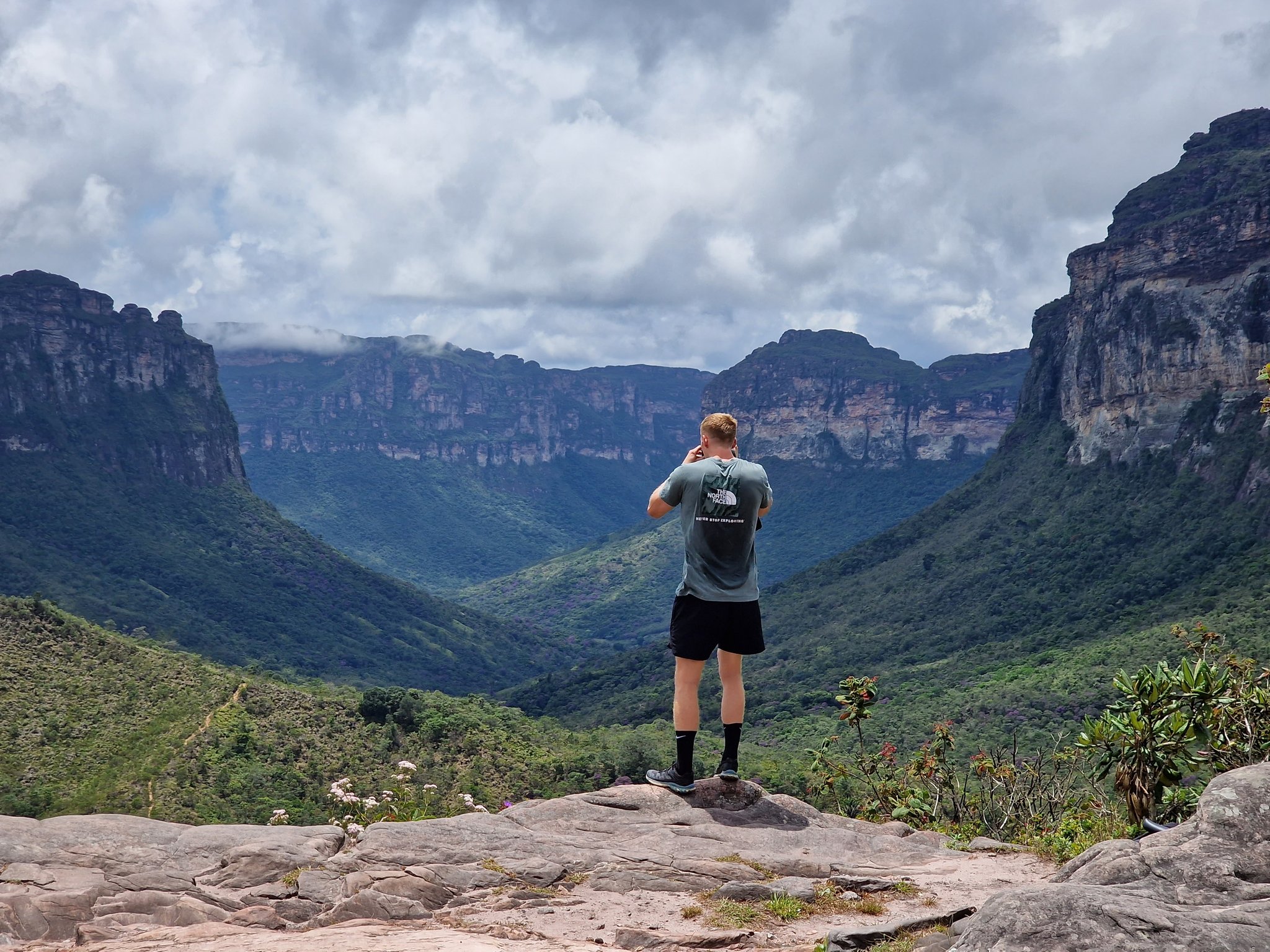 A hiker stands on a rocky outcrop photographing the vast Vale do Pati Trek viewpoint, with towering sandstone cliffs and a lush green valley stretching into the distance under a cloudy sky in Chapada Diamantina