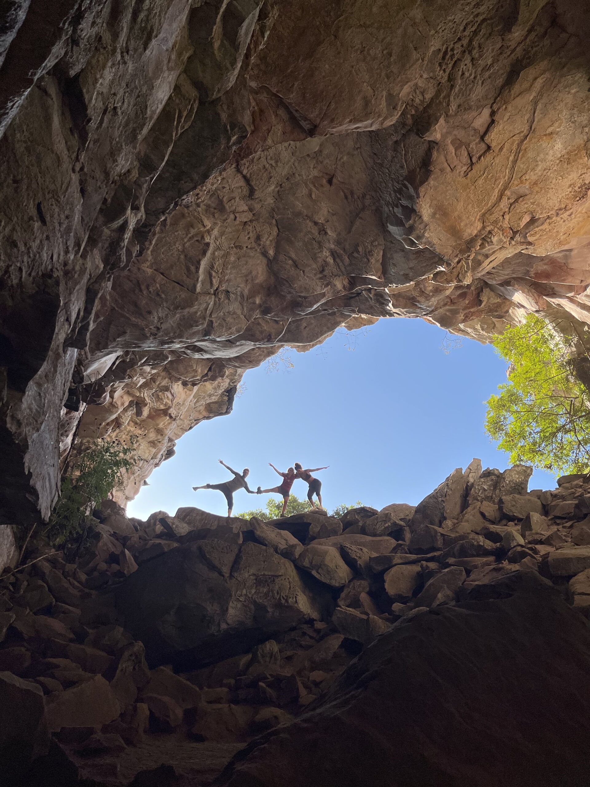 Strike a pose inside the mountain — the cave arch frames everything. Two trekkers striking balance poses on boulders framed by a massive sandstone cave arch on the Vale do Pati Trek, blue sky and green treetops visible beyond the opening