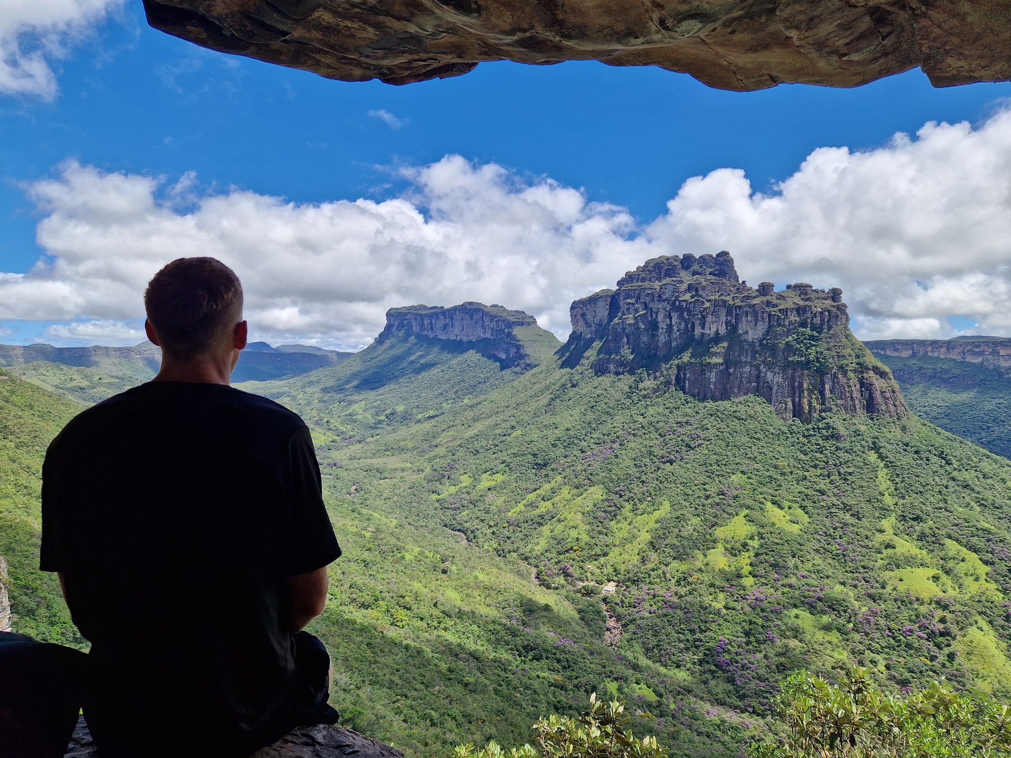 A hiker sits beneath a natural rock overhang gazing out at the dramatic sandstone mesa and lush green valley on the vale do pati trek in Chapada Diamantina, Brazil