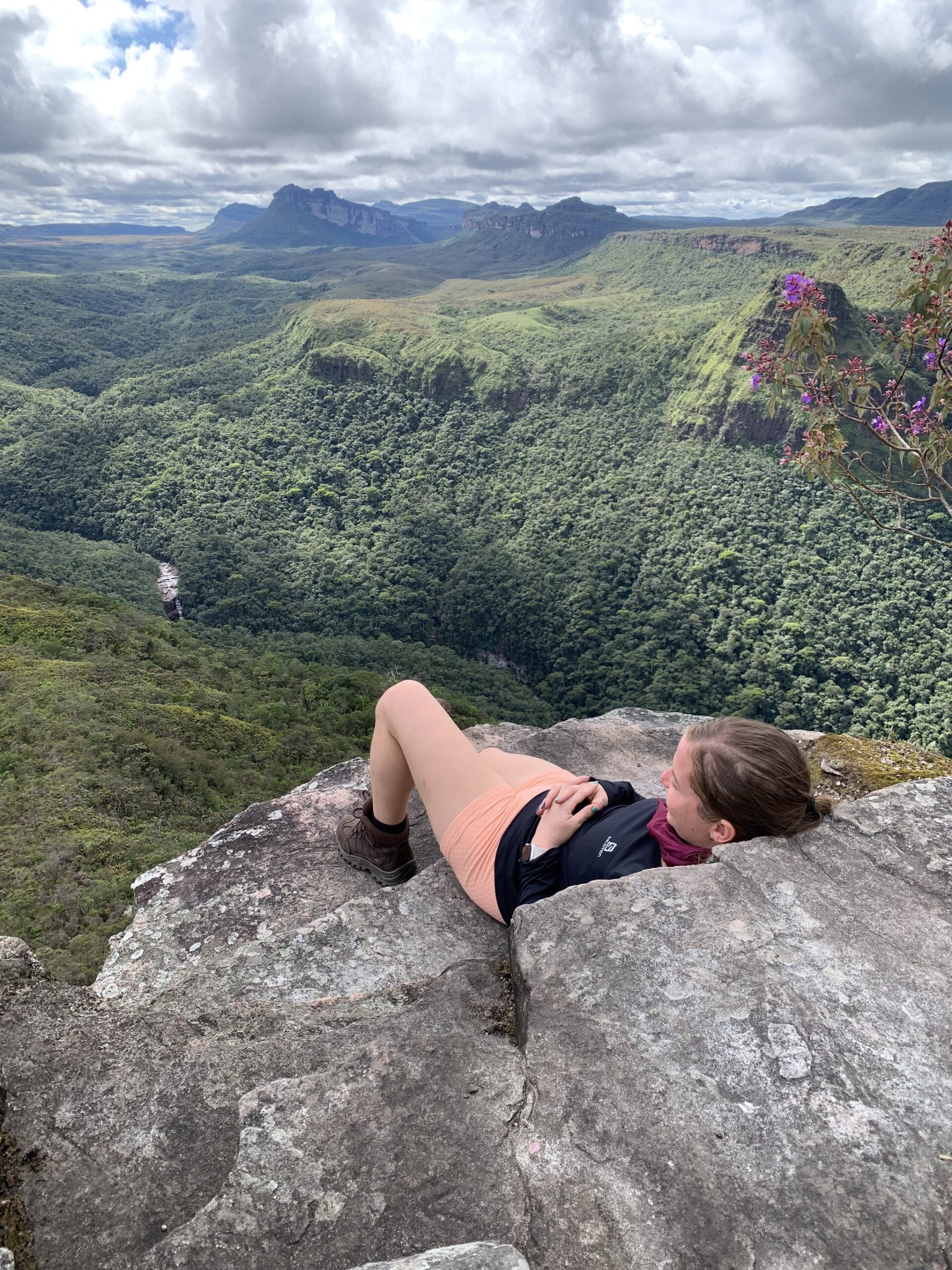 Lie back, breathe deep — the whole valley belongs to you. A hiker lies on a rocky cliff edge on the Vale do Pati Trek, gazing over a vast forested canyon with flat-topped sandstone plateaus and a distant waterfall under a cloudy sky in Chapada Diamantina