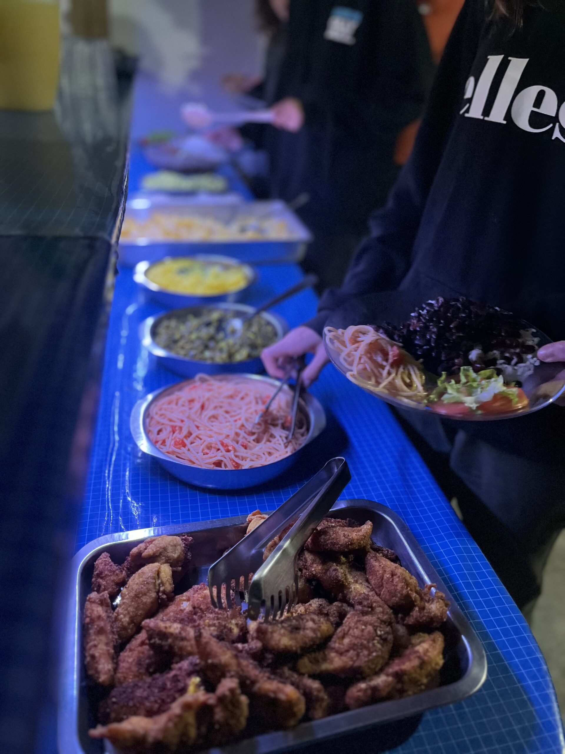 Fuel for the trail — a generous spread of home-cooked food at a family homestay deep in the valley. A long buffet line of home-cooked dishes including breaded fried meat, tomato pasta, lentils, and fresh salad served on a blue-covered table during a Vale do Pati Trek homestay dinner