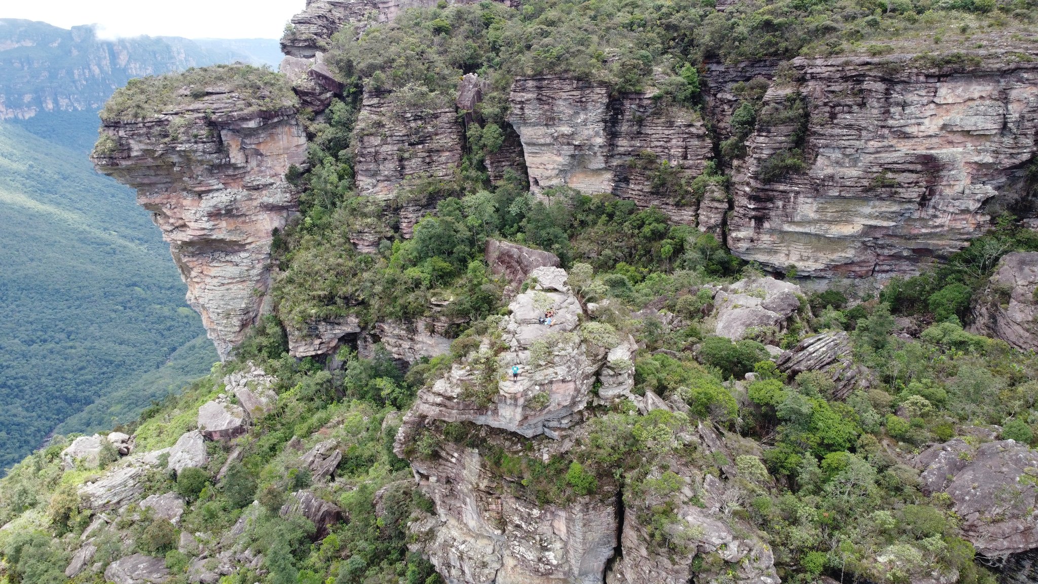 Ancient rock, deep canyon — hikers reduced to specks on the summit. Aerial view of layered sandstone cliffs and green vegetation on the Vale do Pati Trek, with tiny hikers visible on a rocky outcrop