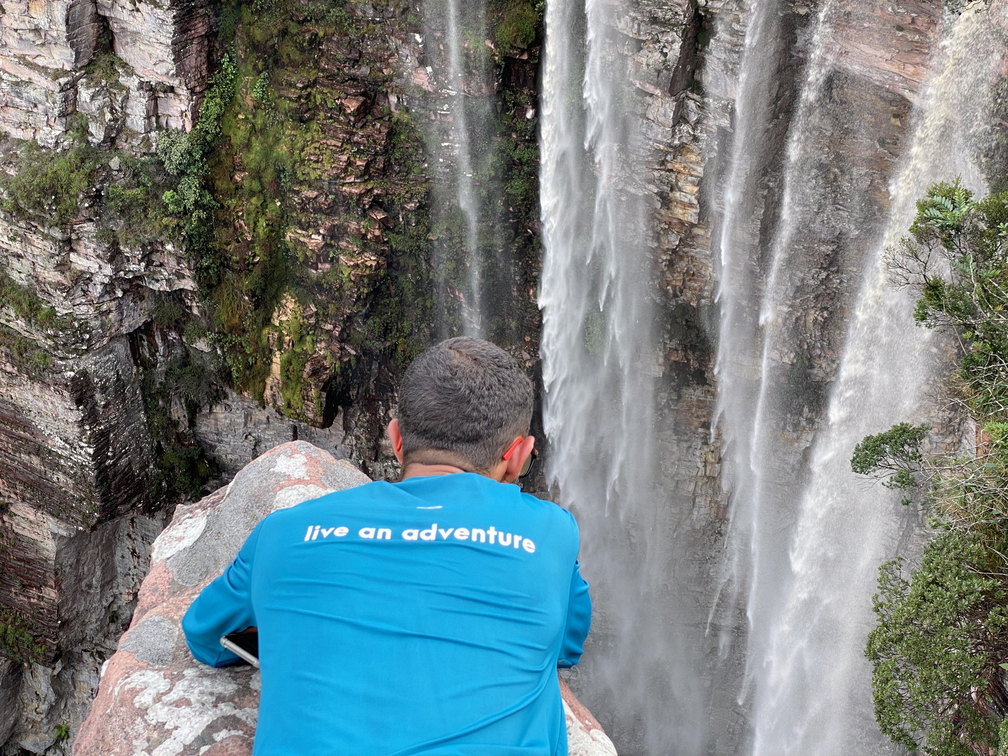 A hiker in a blue 'live an adventure' shirt peers over a boulder toward a powerful multi-strand waterfall cascading down a moss-covered sandstone cliff on the Vale do Pati Trek