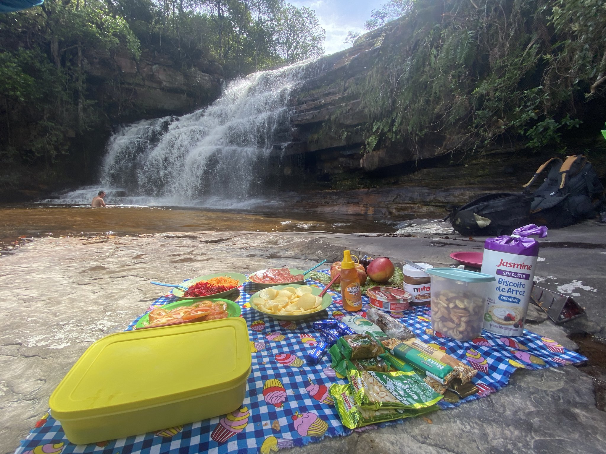 Lunch with a view — a spread of fresh salads, fruit, and snacks right beside a cascading waterfall. A colorful trail picnic laid out on a checkered blanket over flat rocks beside a cascading waterfall on the Vale do Pati Trek, with salads, fruit, nuts, honey, and snacks, while a swimmer enjoys the natural pool in the background