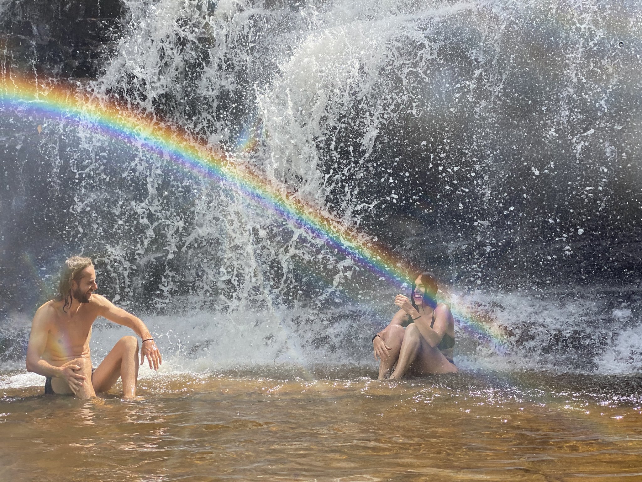 Caught mid-laugh with a rainbow for a backdrop — pure Chapada Diamantina magic. Two smiling travelers sitting in a shallow natural pool at the base of a cascading waterfall on the Vale do Pati Trek, a vivid rainbow arching through the mist between them
