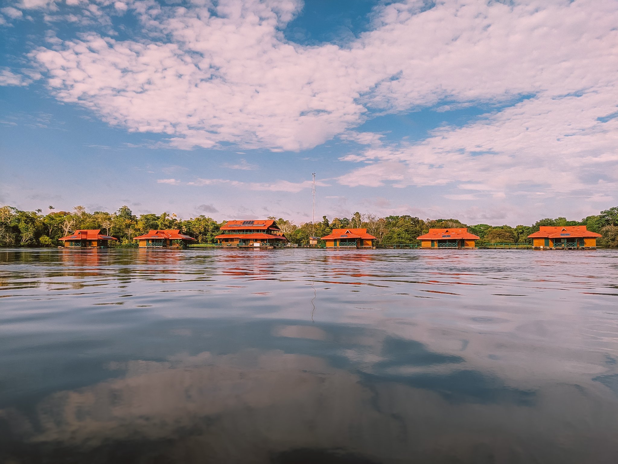 A row of red-roofed wooden bungalows at the Uakari Lodge, an amazon rainforest ecolodge trip in Brazil, seen from water level with clouds reflected in the calm river and dense forest behind